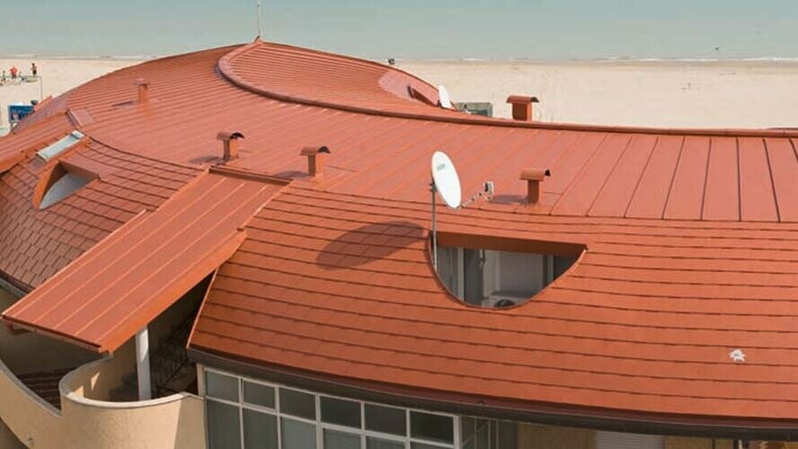 Zaokrąglony dach domu mieszkalnego pokryty dachówką łupkową PREFA oraz dach z rąbkiem stojącym firmy PREFA w kolorze ceglastym.