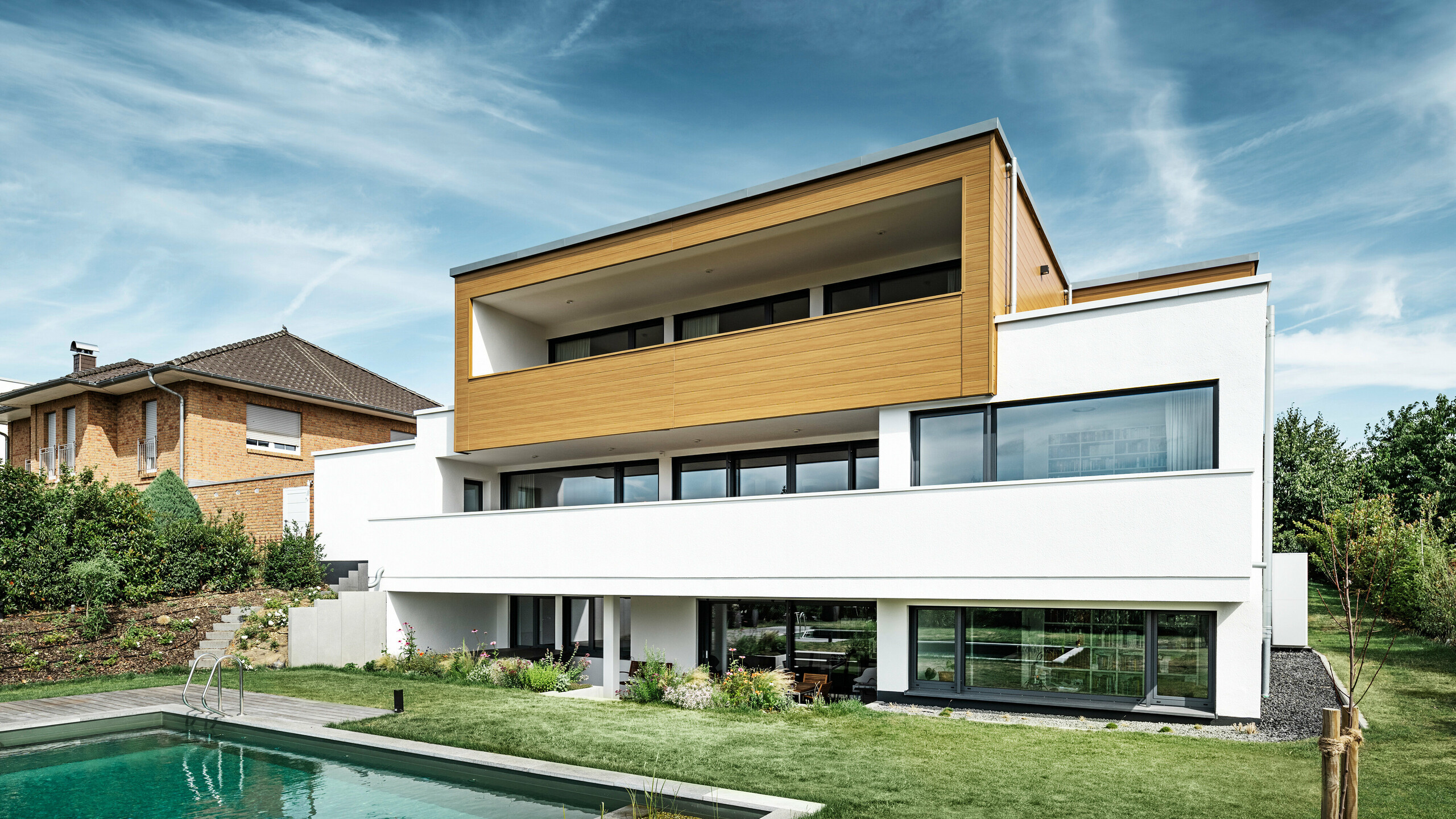 Tył nowoczesnego domu jednorodzinnego w Usingen w Niemczech. W dolnej części zdjęcia widać zielony trawnik, basen i młode drzewo ogrodowe. Przyciągający wzrok dom ma białą podstawę budynku wykonaną z otynkowanej fasady, aluminiową fasadę na piętrze wykonaną z PREFA Sidings w kolorze dąb naturalny i duże powierzchnie okienne z ramą w kolorze antracytu. Sąsiedni dom z ceglaną fasadą jest widoczny z daleka po lewej stronie zdjęcia.