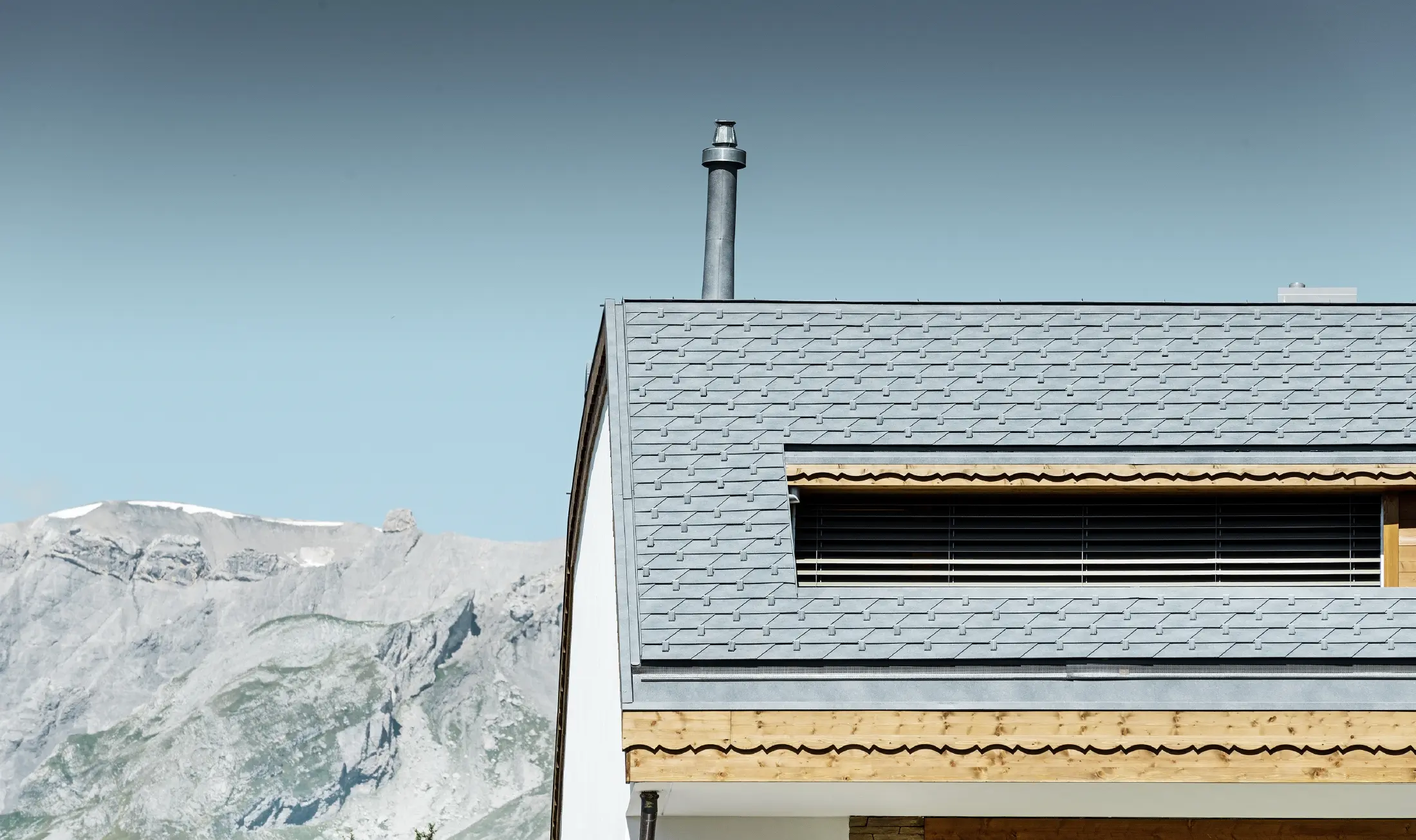Budynek wielorodzinny w Crans-Montana z górami w tle i elewacją z fantazyjnymi elementami drewnianymi skomponowanymi z aluminiową dachówką łupkową PREFA w kolorze kamiennym szarym