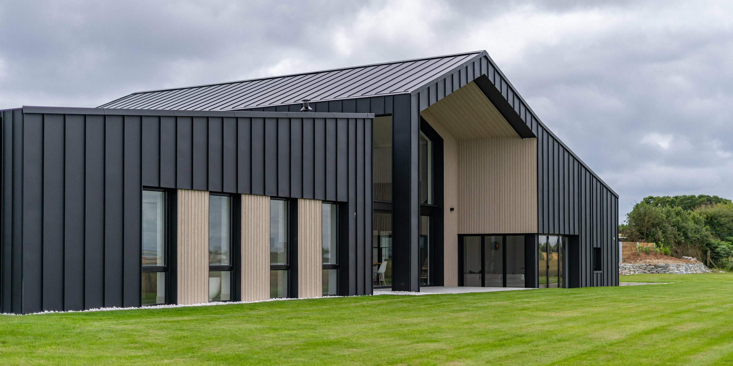 Schrägansicht der Rückseite des Einfamilienhauses ‚The Hide‘ in Cornwall, betont durch die PREFALZ Aluminiumfassade in P.10 Anthrazit. Die präzise Stehfalzdeckung aus hochwertigem Blech bildet einen eleganten Kontrast zu den Holzelementen, während das großzügige Dach mit seiner klaren Linienführung die moderne Architektur des Gebäudes unterstreicht. Umgeben von gepflegtem Grün fügt sich das Design harmonisch in die Landschaft ein.