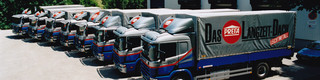 Osiem byłych ciężarówek PREFA w kolorze niebieskim i srebrnym z logiem PREFA stoi w jednym rzędzie