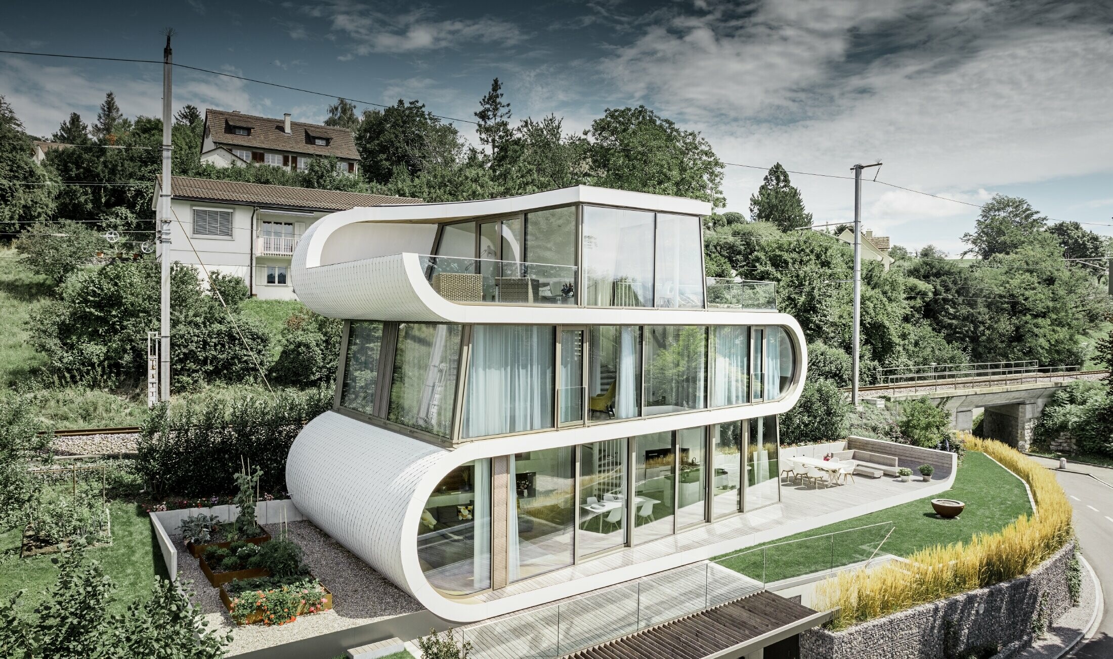Bardzo nowoczesny dom jednorodzinny zaprojektowany przez architekta Camezinda z Zurychu. Wijący się pas łączy ze sobą poszczególne poziomy. Zaokrąglenia zostały obłożone małą dachówką rombem PREFA w kolorze czystym białym. Dom ma wiele dużych powierzchni przeszkolonych, przez co sprawia wrażenie bardzo otwartego.