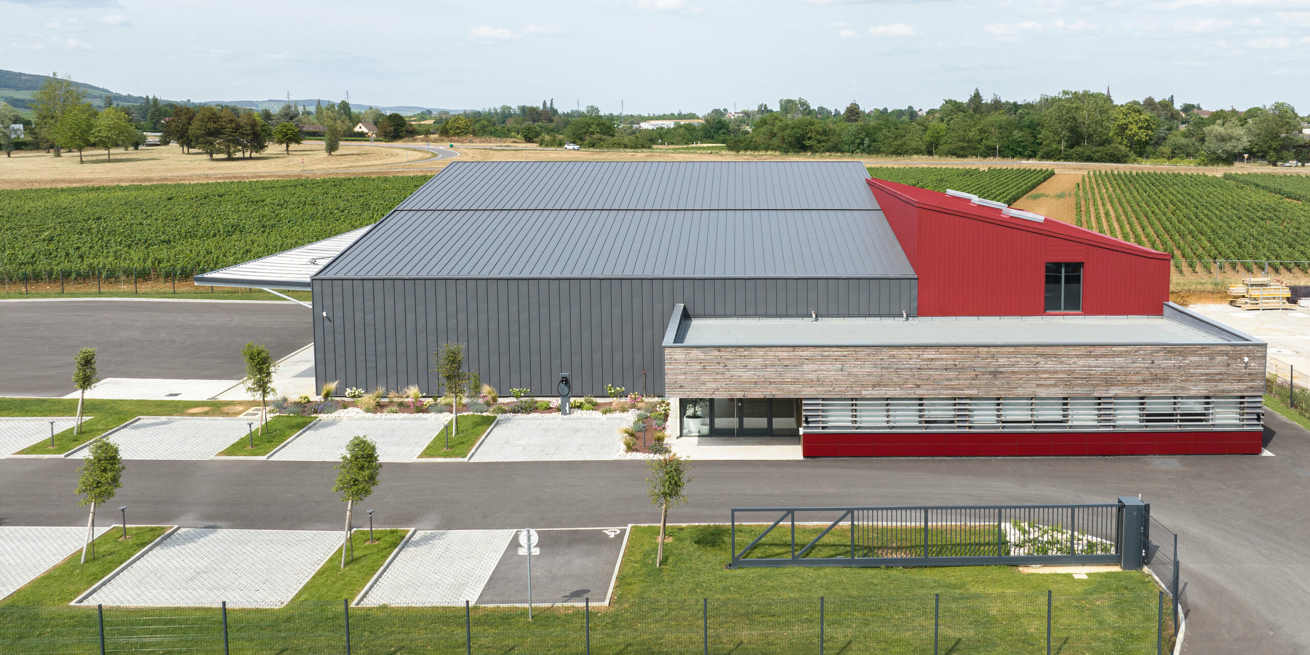 Großes Industriegebäude mit dunkelgrauem PREFALZ am Dach und der Fassade. Als Farbakzent wurden Teile des Gebäudes mit PREFALZ und PREFA Sodings in P.10 Oxydrot verkleidet. Das Gebäude ist zur Landschaft mit Feldern im Hintergrund ausgerichtet. Im Vordergrund des Bildes sind Parkplätze mit jungen Bäumen und gepflegter Bepflanzung zu sehen.