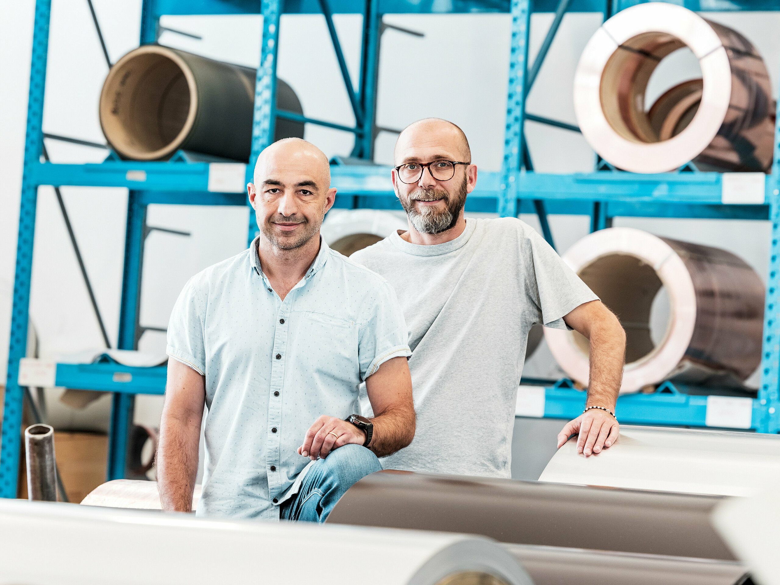 Dwóch rzemieślników w hali z aluminium przed nawiniętymi metalowymi szpulami – nowoczesne środowisko pracy dla aluminiowych systemów dachowych i elewacyjnych.