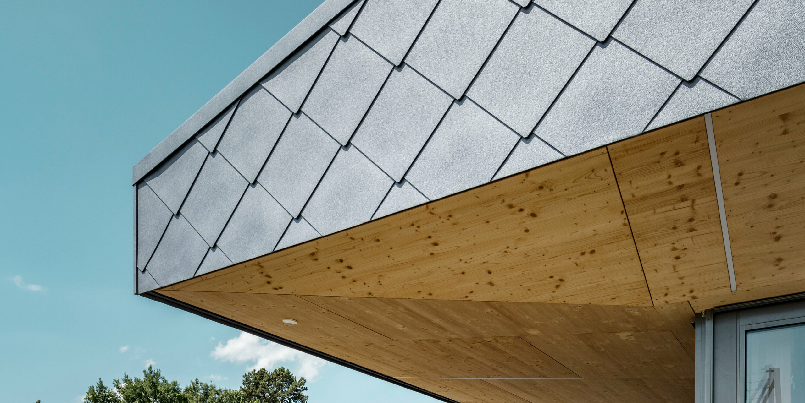 Detailaufnahme des innovativen Dachübergangs einer Sportanlage in Prilly, Schweiz, mit PREFA Dach- und Wandrauten 44x44 in P.10 Steingrau. Die Rauten aus Aluminium bieten eine ästhetische und funktionale Bedeckung, die nahtlos in die warme, natürliche Holzuntersicht übergeht. Die Kombination von langlebigen Aluminiumelementen mit dem natürlichen Holzakzent zeugt von einem architektonischen Ansatz, der Nachhaltigkeit und modernes Design vereint.