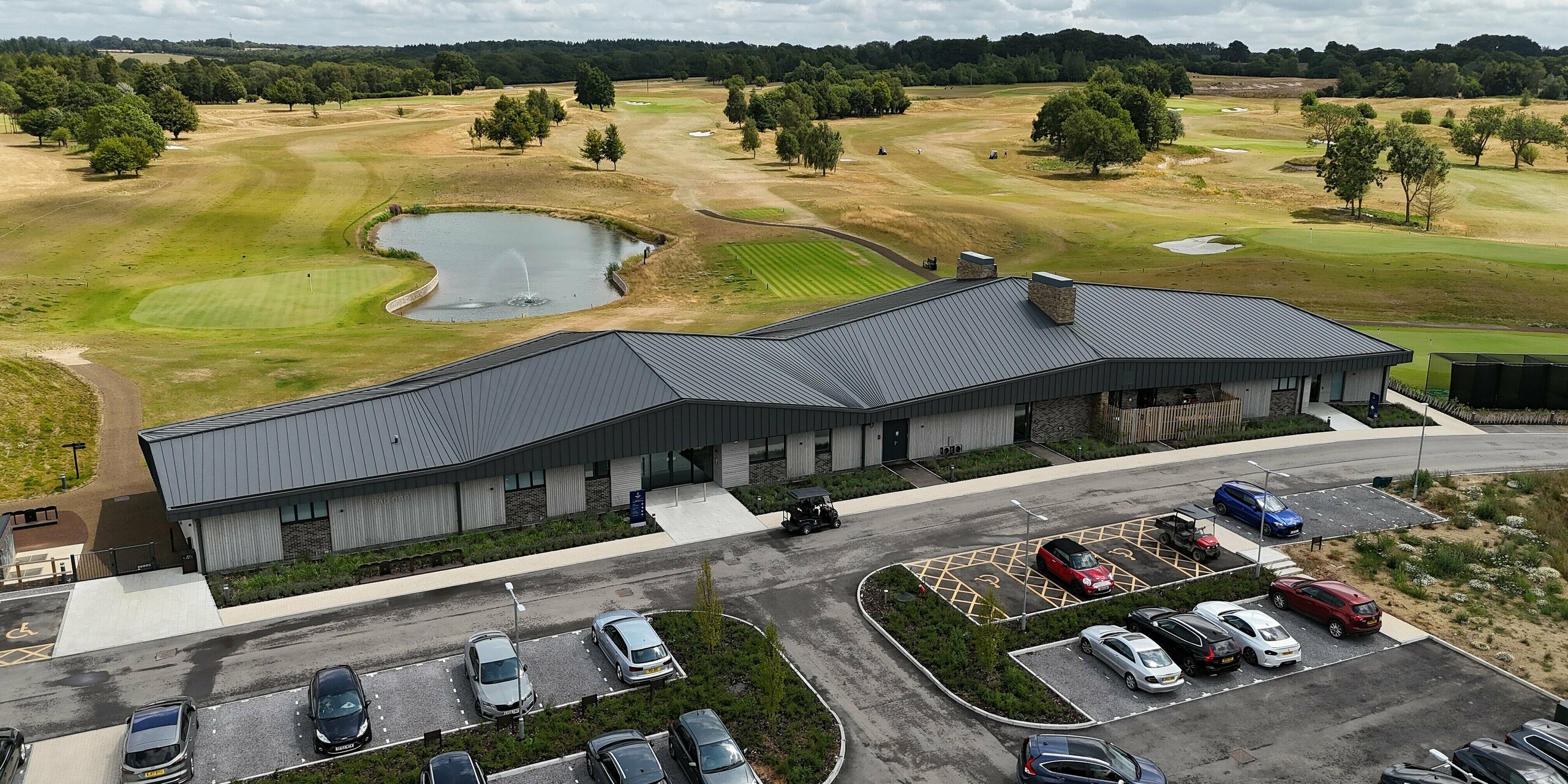 Der moderne Neubau des Basingstoke Golf Clubs in Hampshire beeindruckt mit einem markanten PREFALZ Stehfalz-Dach in P.10 Dunkelgrau. Die dynamische Dachform und die stilvolle Fassade aus Holz und Stein fügen sich perfekt in die weitläufige Golfplatzlandschaft ein. Das Gebäude liegt malerisch am Wasser, umgeben von gepflegten Grünflächen. Im Vordergrund ist der Parkplatz mit Fahrzeugen und einem Golfcart zu sehen. Das widerstandsfähige, wartungsarme Aluminium des Daches sorgt für langanhaltenden Schutz vor Witterungseinflüssen und verleiht dem Clubhaus eine zeitgemäße, funktionale Ästhetik.