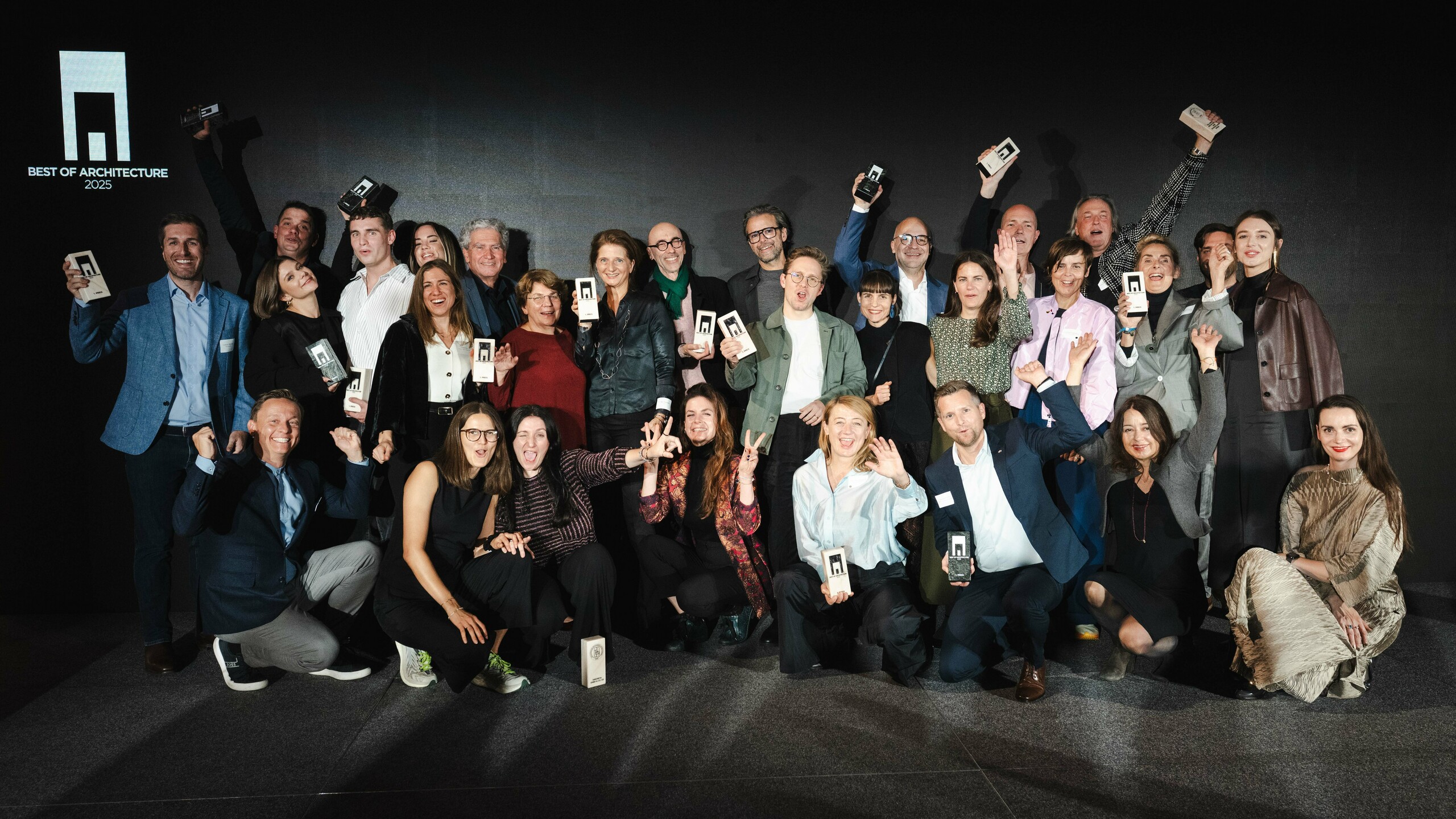 Zdjęcie grupowe zwycięzców podczas ceremonii wręczenia nagród „Budynek mieszkalny roku 2025”: zwycięzcy stoją przed elegancką czarną sceną z logo „Best of Architecture” i dumnie trzymają w rękach swoje nagrody. Dynamiczne pozy, nowoczesne ubrania i efektowne oświetlenie podkreślają znaczenie wydarzenia, a scena symbolizuje innowacyjność i doskonałość architektoniczną.