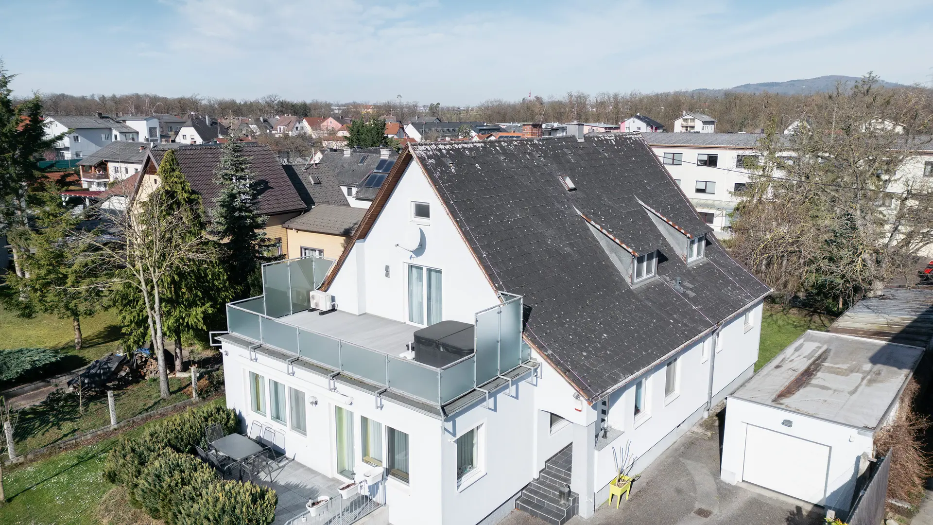 Widok z lotu ptaka na budynek ze starym dachem z eternitu, pokrytym brudem i porostami. Na dachu nie widać żadnych paneli słonecznych. Balkon na pierwszym planie ma szklaną balustradę i znajduje się na najwyższym piętrze budynku.