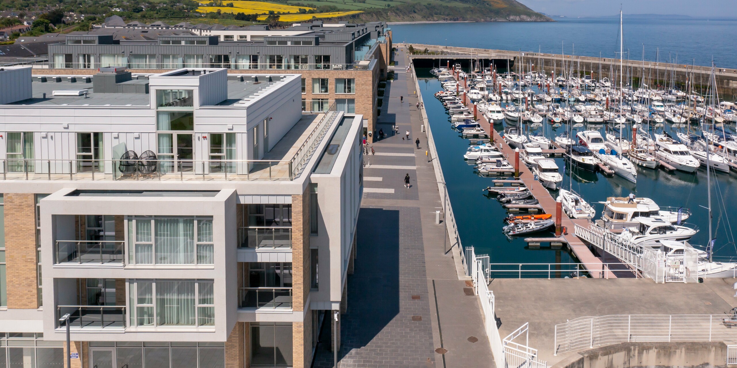 Luftaufnahme von Marina Village Greystones in Wicklow, Irland – Moderne Wohnsiedlung mit direkter Lage am Yachthafen. Die Fassade des Wohnkomplexes ist mit ca. 2.000 m² robustem PREFALZ in den Farben P.10 Prefaweiß und P.10 Hellgrau gestaltet. Elegante Architektur mit großzügigen Glasfronten und Balkonen, umgeben von malerischer Landschaft am Wasser. Die hochwertige Aluminiumverkleidung von PREFA bietet dauerhaften Schutz und zeitlose Ästhetik.