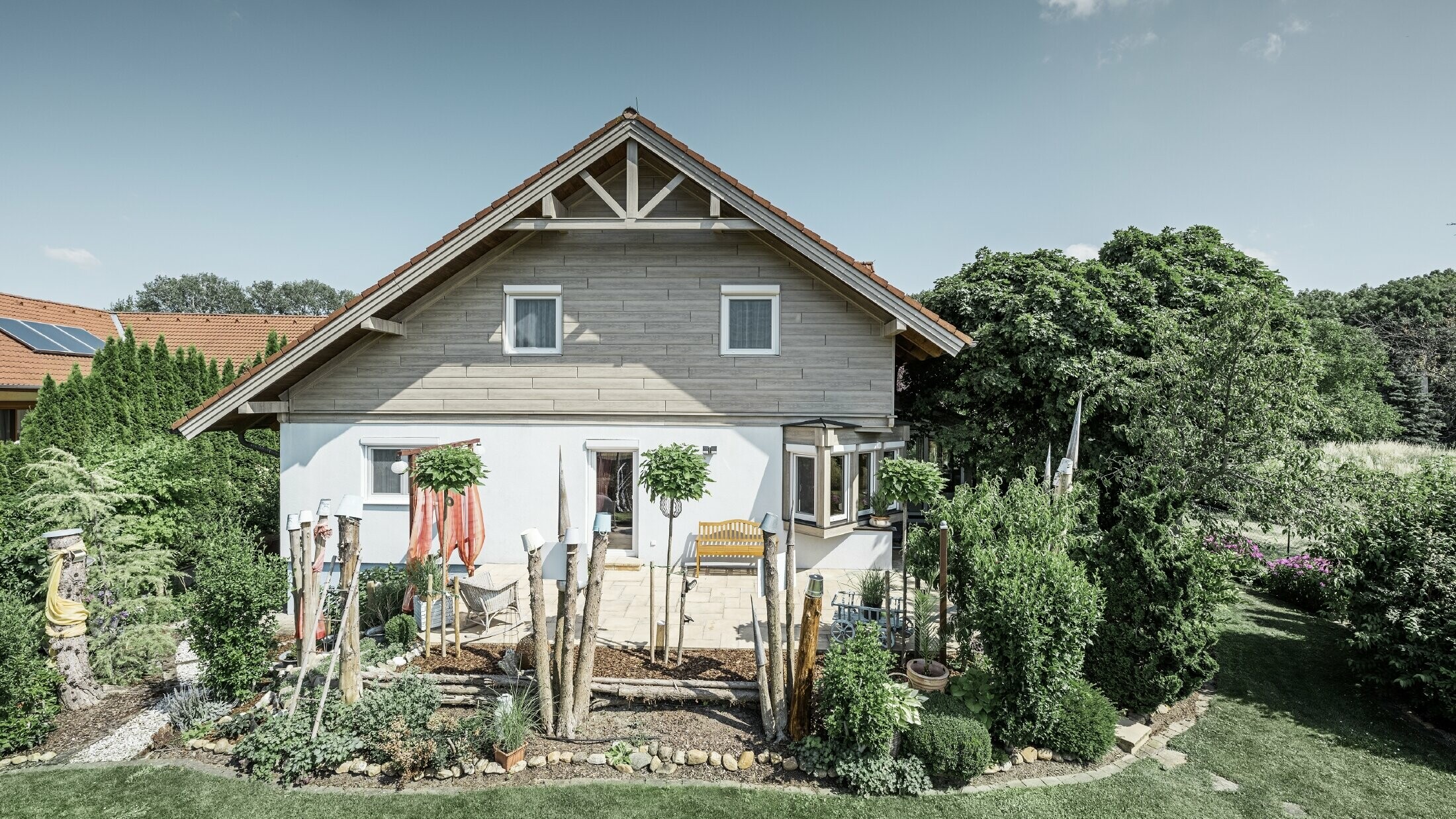 Dom jednorodzinny z odnowioną elewacją i nową okładziną szczytową w nowoczesnym stylu imitującym drewno w odcieniach szarych i beżowych, w połączeniu z elewacją otynkowaną, dachem dwuspadowym i zadbanym ogrodem.