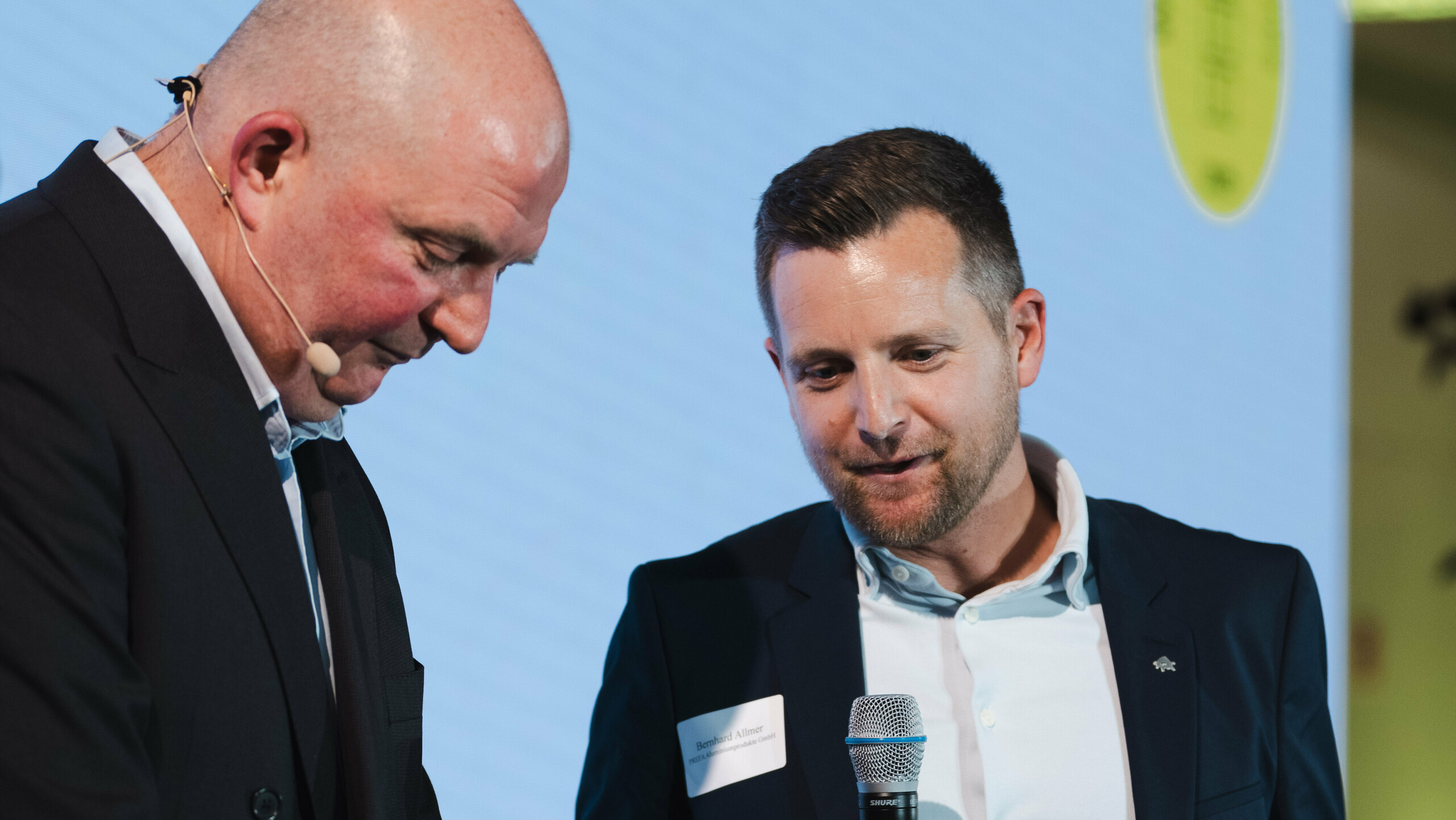 Wręczenie nagród w konkursie „Budynek mieszkalny roku” 2025: Na scenie stoi dwie osoby, jedna trzyma mikrofon i nagrodę „Architects' Choice”, a druga przygotowuje się do jej wręczenia. Mężczyzna z mikrofonem i nagrodą to Bernhard Allmer, odpowiedzialny za produkty solarne PREFA. W tle widoczna jest duża powierzchnia projekcyjna z logo wydarzenia i zielonymi akcentami. Scena symbolizuje innowacyjność, jakość architektoniczną i uznanie modułu solarnego PREFALZ jako zrównoważonego rozwiązania dla nowoczesnych systemów dachowych.