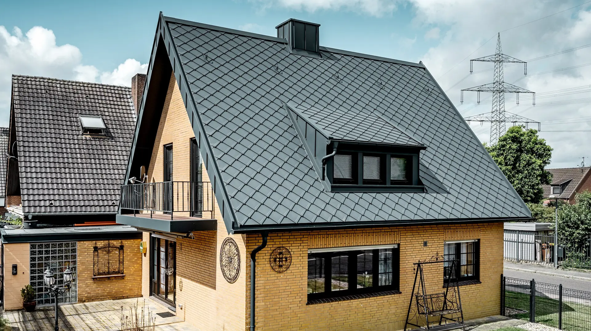 Dom jednorodzinny z żółtej cegły klinkierowej po renowacji dachu z dachówką PREFA romb 29 × 29 w kolorze grafitowym; nowo pokryty, stromy dach dwuspadowy z wzorem rombowym, zintegrowaną lukarną i kominem, tarasem i ogrodem na pierwszym planie.