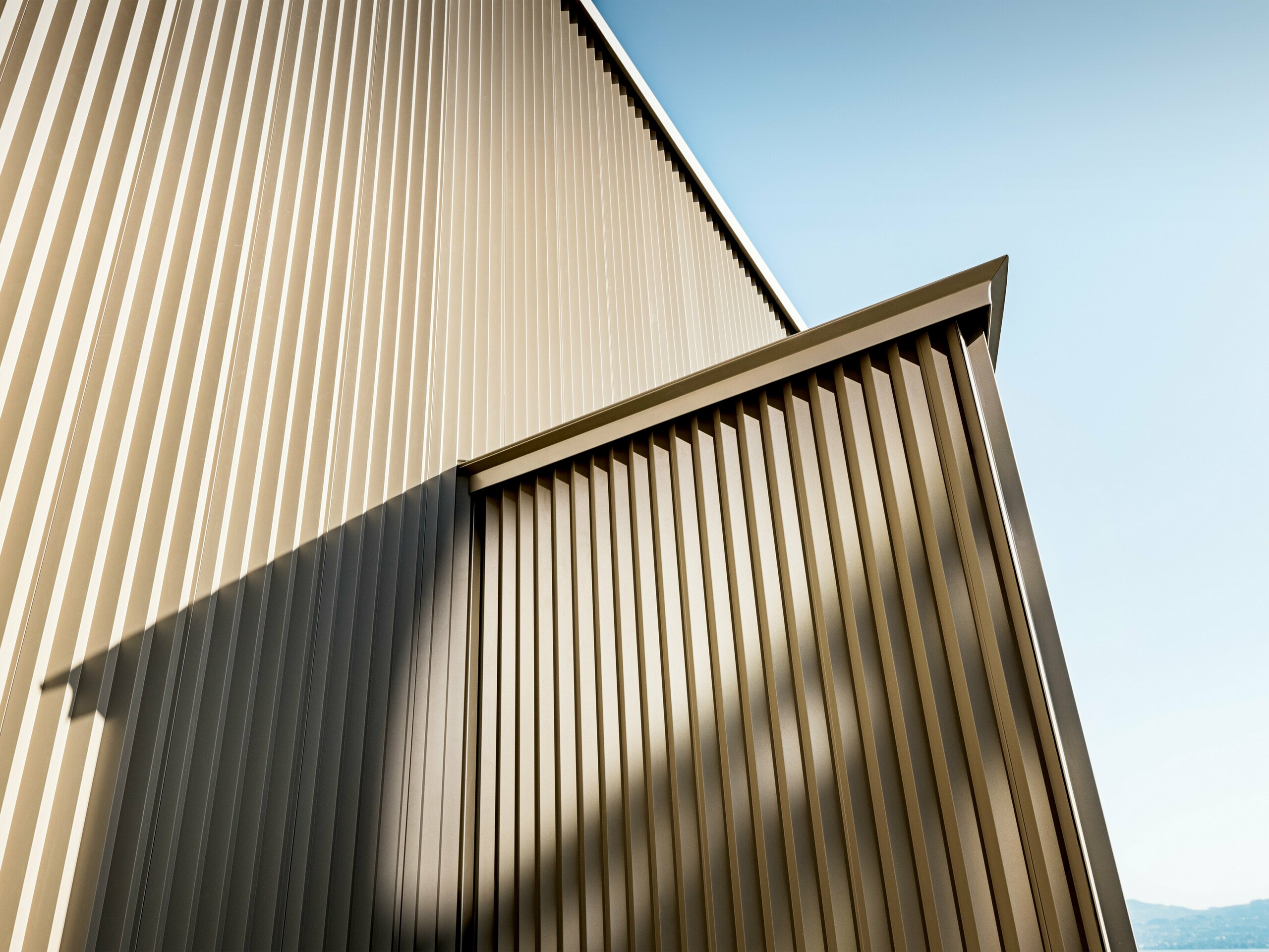 Elegancka fasada aluminiowa PREFA w kolorze brązu na budynku mieszkalnym w St. Gingolph, zaprojektowana przez architektkę Sophie Morard. Pionowe, postrzępione profile tworzą fascynującą grę światła, cienia i struktury, nadając elewacji głębię i dynamikę. Wysokiej jakości powierzchnia o powłokach proszkowych zapewnia długotrwałą ochronę i stylowy wygląd – idealny dla wymagających projektów mieszkaniowych. Brązowy połysk harmonijnie komponuje się z alpejskim otoczeniem i sprawia, że ten niewielki domek staje się architektoniczną atrakcją o dużym efekcie.