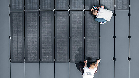 Dwie osoby montują moduły solarne PREFALZ na dużej, szarej powierzchni dachu, wyłożonej pasami ze stojącym rąbkiem podwójnym.