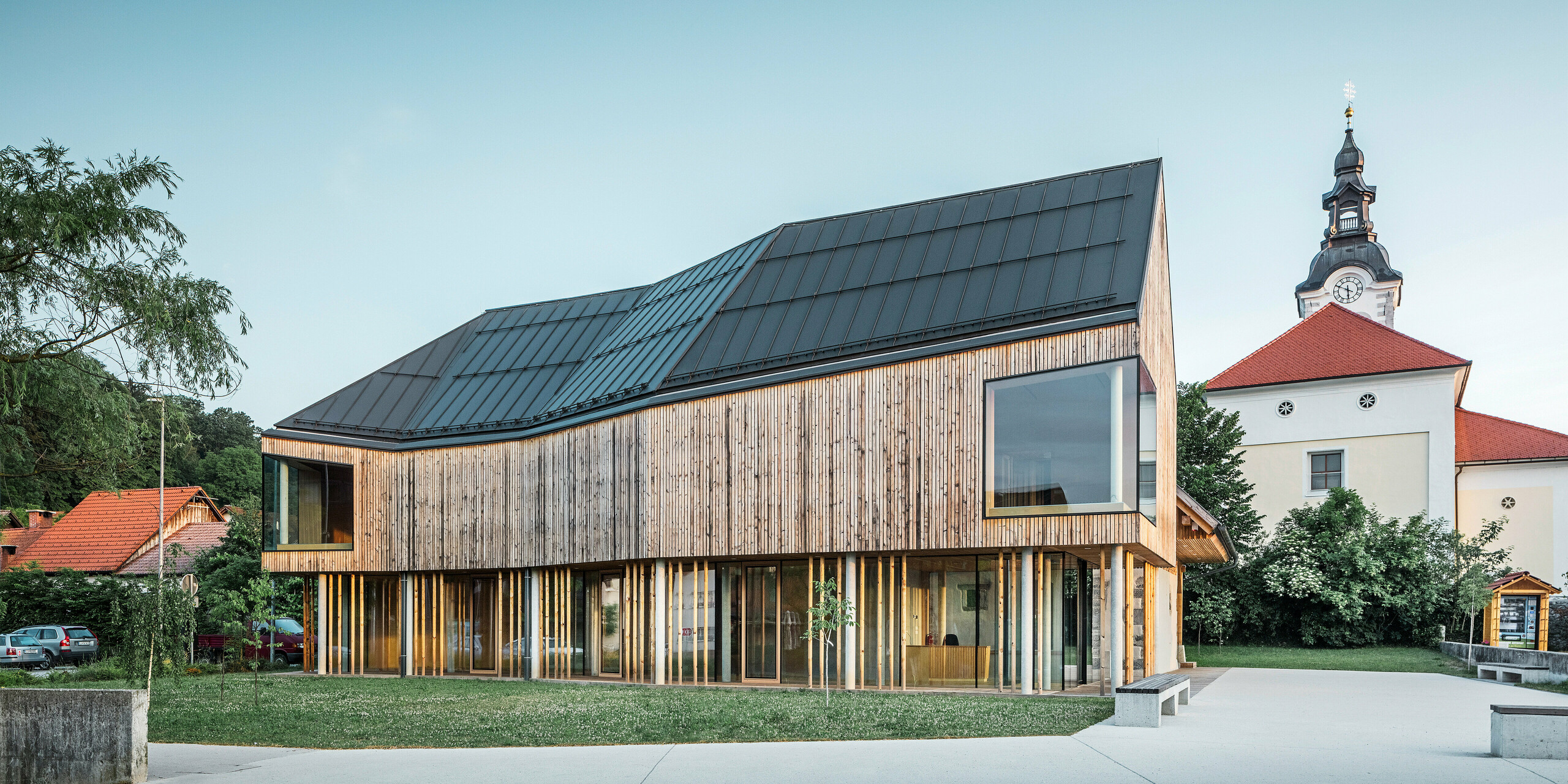 Das Besucherzentrum in Ig in Slowenien zeigt eine elegante Kombination aus traditioneller und moderner Architektur. Das langlebige PREFALZ Dachsystem in P.10 Dunkelgrau harmoniert stilvoll mit der Holzfassade und dem historischen Kirchturm im Hintergrund. Die großen Fenster spiegeln den blauen Himmel wider und betonen die Transparenz und Offenheit des Entwurfs, der die kulturelle Bedeutung des Zentrums als Bildungs- und Informationszentrum widerspiegelt. Ein Beispiel für die zeitgenössische Dachgestaltung von PREFA.