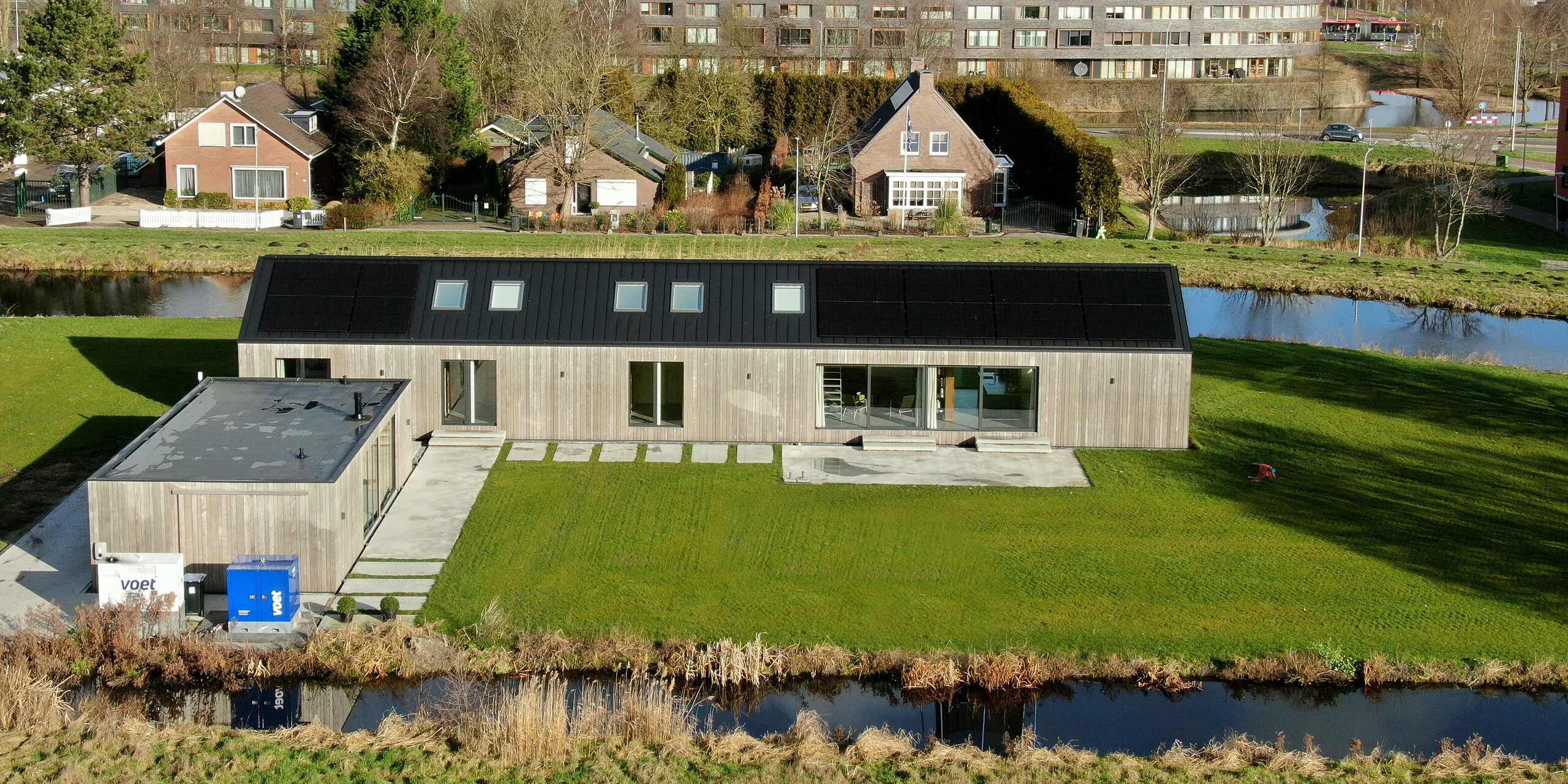 Vogelperspektive auf einen modernen Bungalow inkl. Nebengebäude in Uithoorn, Niederlande, mit Winkelstehfalz-Eindeckung aus schwarzgrauem PREFALZ und integrierter Photovoltaik-Anlage. Das Bild zeigt die gesamte Anlage mit großzügigem Garten und umgebendem Wassergraben. Das Haus zeichnet sich durch große Glasfronten und eine natürliche Holzfassade aus, die eine harmonische Verbindung zur Umgebung schaffen. Ideal für nachhaltige Bauprojekte, die erneuerbare Energien und modernes Design integrieren.