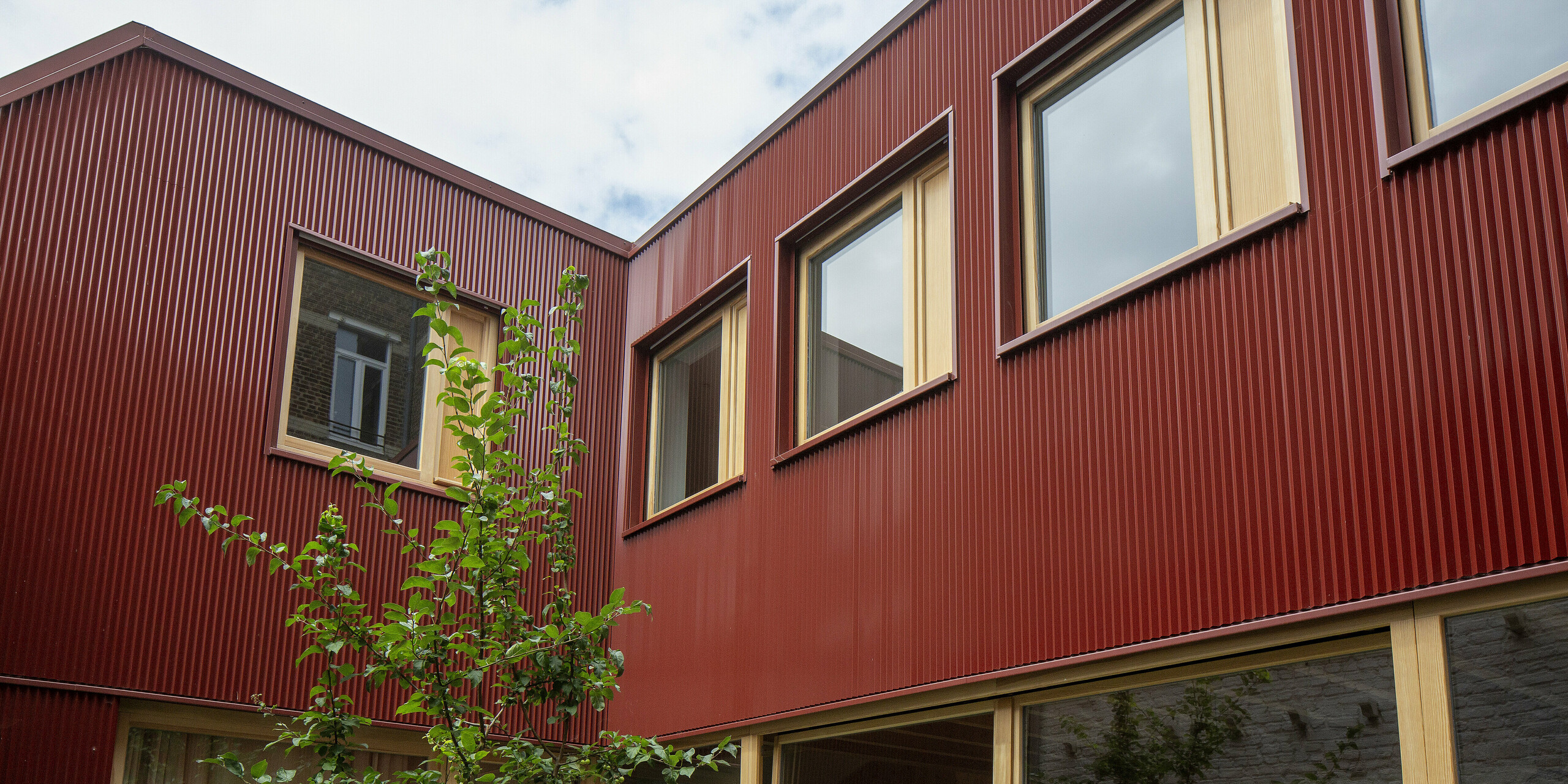 Der rote Innenhof in Lille beeindruckt durch das lebendige Zusammenspiel von vertikalem PREFA-Zackenprofil, warmem Holz und großflächigen Fenstern. Die kräftige Farbe Oxydrot bildet einen harmonischen Kontrast zum frischen Grün der Pflanzen und ist ein langlebiger, wartungsfreier Blickfang mit hoher architektonischer Wirkung.