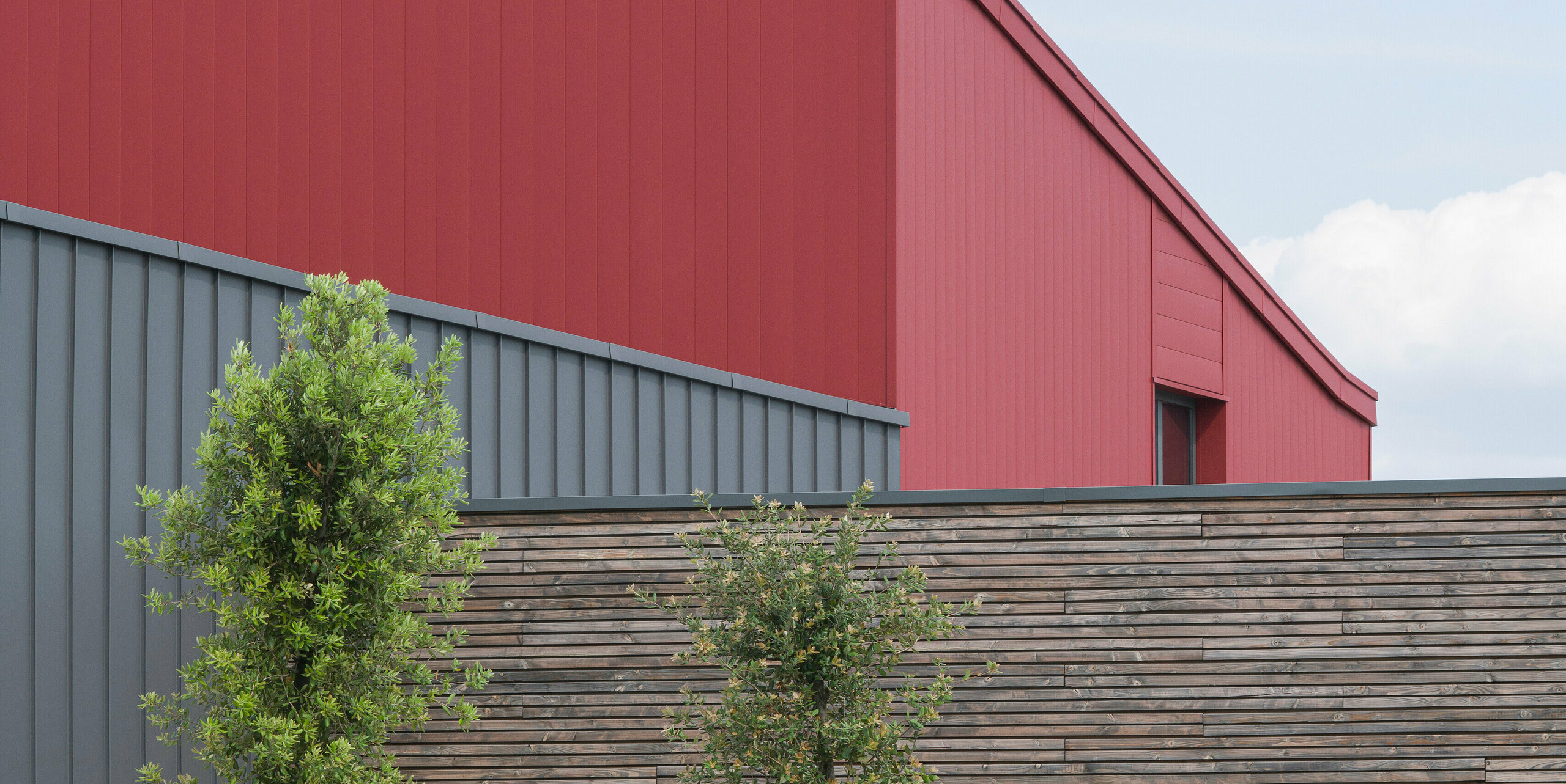 Ein modernes Geschäftshaus mit einer auffälligen roten Aluminiumfassade. Die Wahl der Farbe spricht eine klare architektonische Sprache. Die strukturierte, graue Aluminiumverkleidung bildet einen subtilen Kontrast zum markanten Rot. Die langlebige PREFALZ Aluminiumverkleidung an Fassade und Dach bietet idealen Schutz gegen alle Witterungsbedingungen. Die ergänzende Naturholzverkleidung vermittelt Wärme und Natürlichkeit. Vor dem Gebäude sind junge Bäume zu sehen, die sorgfältig mit Stützpfosten gesichert sind. Ein gut gepflegtes Blumenbeet sorgt für zusätzliche Farbe und Leben. Eine Ladestation für Elektroautos im Vordergrund zeigt das Engagement für nachhaltige Energielösungen.