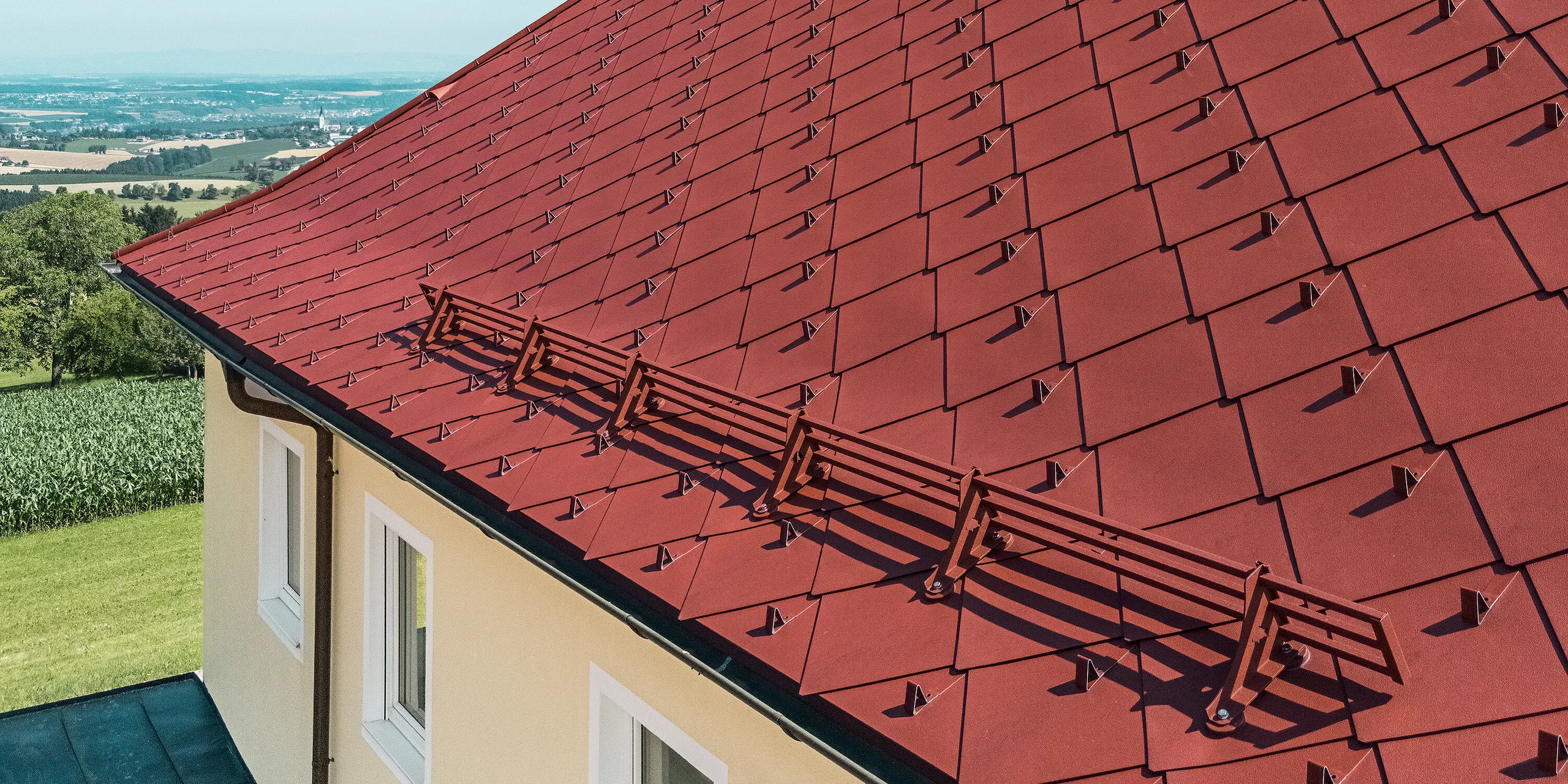 Das Bild zeigt das Dach eines sanierten Einfamilienhauses in Aschach an der Steyr, das mit der PREFA Dachraute 44 x 44 in der Farbe P.10 Oxydrot ausgestattet ist. Die markante rote Farbe des Daches fügt sich harmonisch in die umgebende Natur ein. Besonders auffällig ist der Schneefang aus Metall, der für Dach-Sicherheit sorgt. Zusätzlich wurden die einzelnen Dachrauten mit Schneestoppern ausgestattet, um das Abrutschen von Dachlawinen zu verhindern.