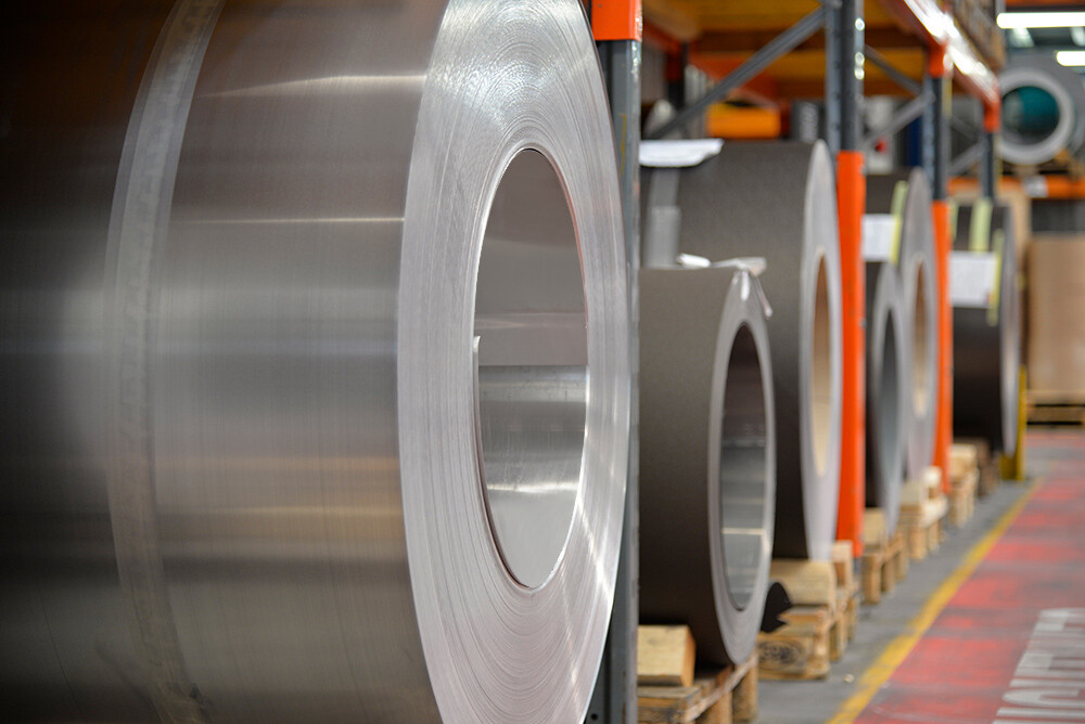 Aluminiumrollen (Coils) gelagert auf Holzpaletten, im Vordergrund steht eine naturblanke Alurolle, dahinter findet man weitere Bänder und Bleche aus Aluminium.