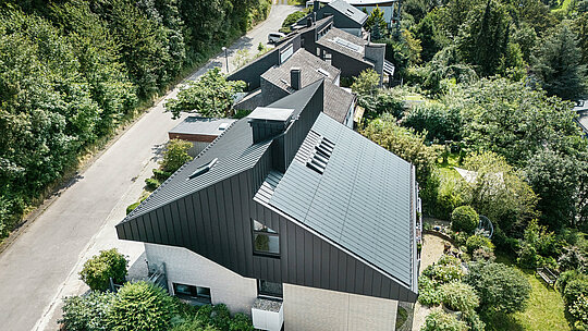 Widok z boku nowoczesnego domu jednorodzinnego w Meschede w Niemczech. Dach dwuspadowy pokryty jest blachą PREFALZ P.10 czarny. Aluminiowy dach stanowi atrakcyjny akcent wizualny i zapewnia budynkowi długotrwałą ochronę. Precyzyjna konstrukcja podwójnego dachu rozciąga się od dachu przez elewację aż do przodu budynku. Otoczony bujną zielenią i spokojną ulicą, energooszczędny dom z innowacyjnymi modułami solarnymi PREFALZ harmonijnie wtapia się w naturalne otoczenie.