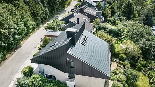Widok z boku nowoczesnego domu jednorodzinnego w Meschede w Niemczech. Dach dwuspadowy pokryty jest blachą PREFALZ P.10 czarny. Aluminiowy dach stanowi atrakcyjny akcent wizualny i zapewnia budynkowi długotrwałą ochronę. Precyzyjna konstrukcja podwójnego dachu rozciąga się od dachu przez elewację aż do przodu budynku. Otoczony bujną zielenią i spokojną ulicą, energooszczędny dom z innowacyjnymi modułami solarnymi PREFALZ harmonijnie wtapia się w naturalne otoczenie.