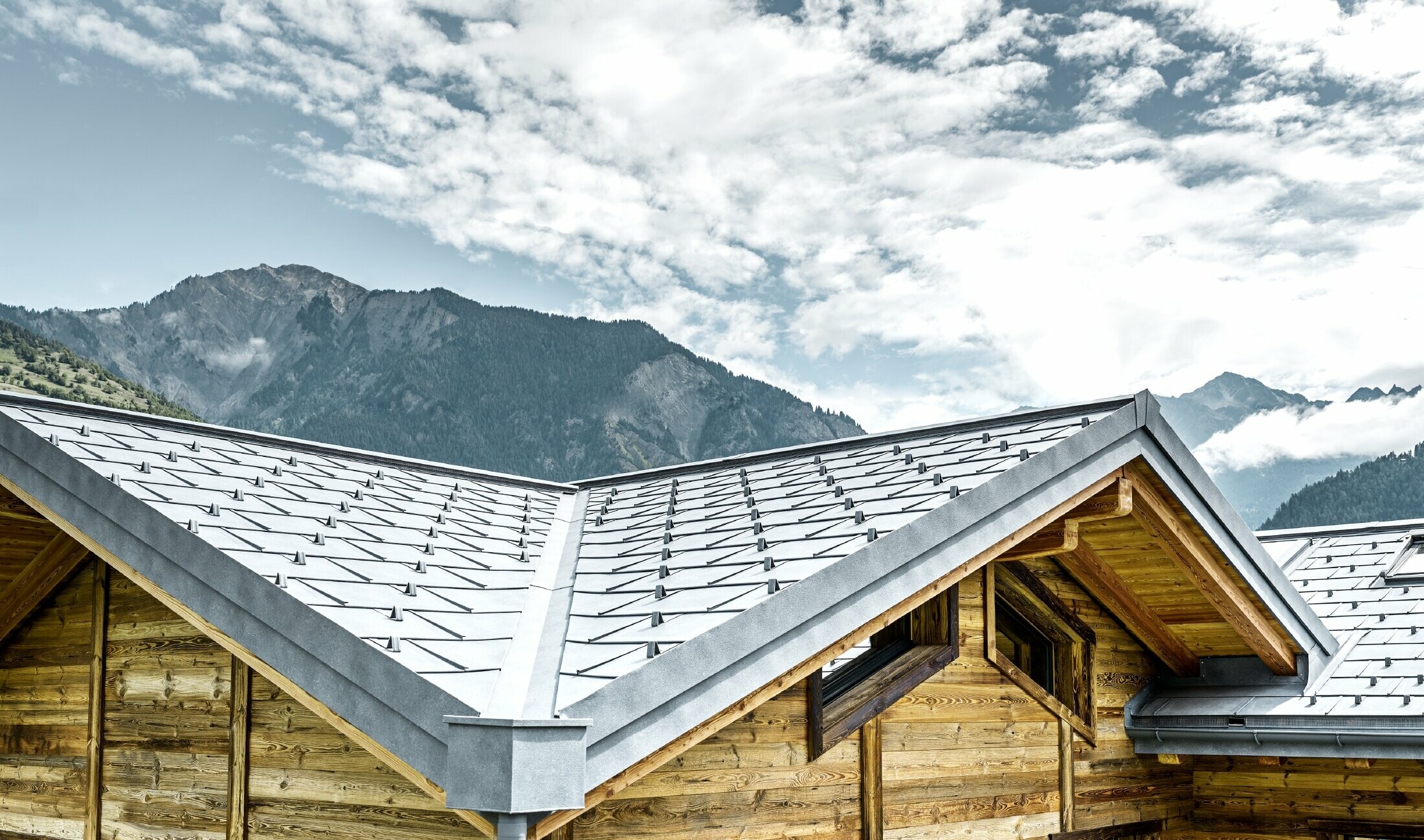 Dach rustykalnej chaty w Szwajcarii z drewnianą elewacją i aluminiowym dachem PREFA. Wykorzystana została dachówka klasyczna R.16 PREFA w kolorze szarym kamiennym. Widać strukturę dachu i wgłębienie.