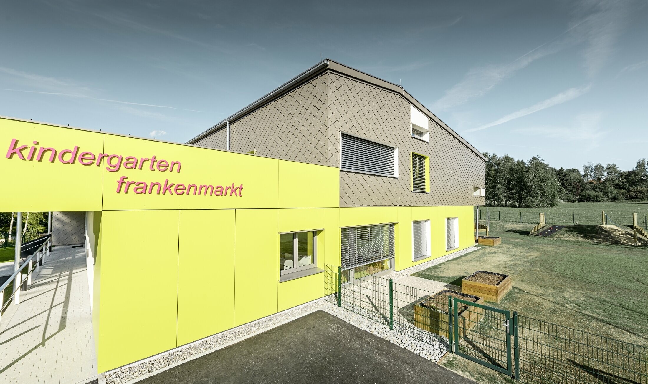 Elewacja przedszkola we Frankenmarkt została częściowo obłożona rombem fasadowym PREFA w kolorze brązowym. Resztę elewacji obłożono żółtymi płytami elewacyjnymi. Widoczny jest także napis z nazwą przedszkola.