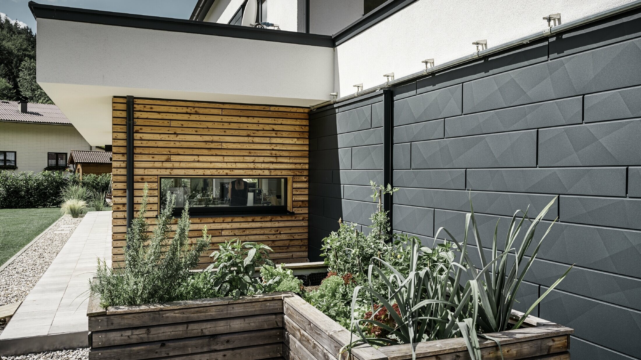 Zdjęcie przedstawia zbliżenie domu jednorodzinnego, na którym uwaga skupia się na okładzinie elewacyjnej PREFA Siding.X w kolorze P.10 antracyt. Ciemne panele elewacyjne o charakterystycznym, pofałdowanym wyglądzie tworzą atrakcyjny kontrast wizualny z ożywionymi, pokrytych drewnem fragmentami oraz otaczającymi terenami zielonymi. Przed elewacją z aluminium znajdują się podwyższone grządki, obsadzone różnymi roślinami i krzewami, co podkreśla naturalny i zrównoważony charakter domu. Połączenie nowoczesnych materiałów i integracja z ogrodem tworzy obraz współczesnej architektury, która harmonijnie wtapia się w otoczenie.