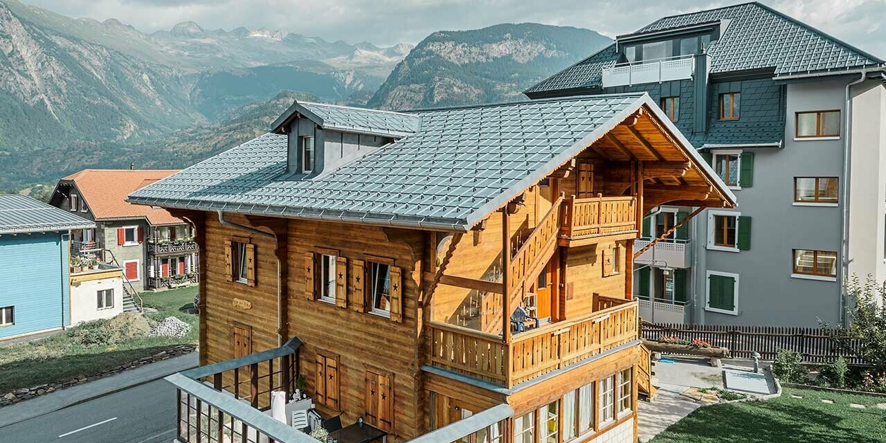 Tradycyjny szwajcarski drewniany domek letniskowy z lukarną i dachem dwuspadowym. Dach jest kryty klasycznymi dachówkami PREFA w kolorze szarym kamiennym.