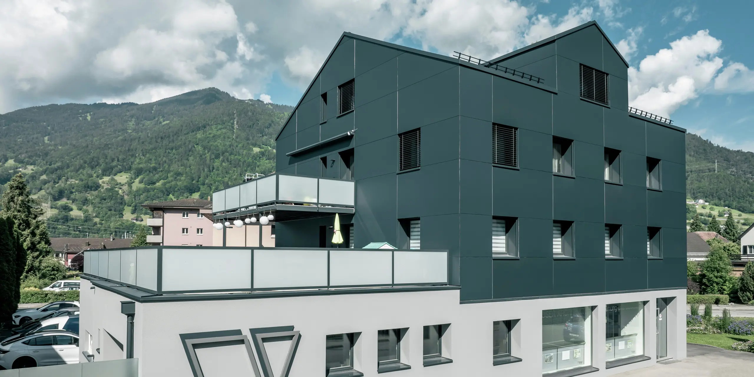 Das Architekturbüro Voser in Landquart, im Schweizer Graubünden, mit einer PREFA Aluminiumfassade in Anthrazit. Das Gebäude wurde saniert und mit PREFABOND Aluminium Verbundplatten ausgestattet. Die Gestaltung kombiniert klare Linien und hochwertige Materialien. Auffällig sind die großen Balkone mit Glasbrüstungen und das prägnante Logo des Architekturbüros Voser an der Gebäudeseite. Im Hintergrund ist eine grüne Berglandschaft zu erkennen.