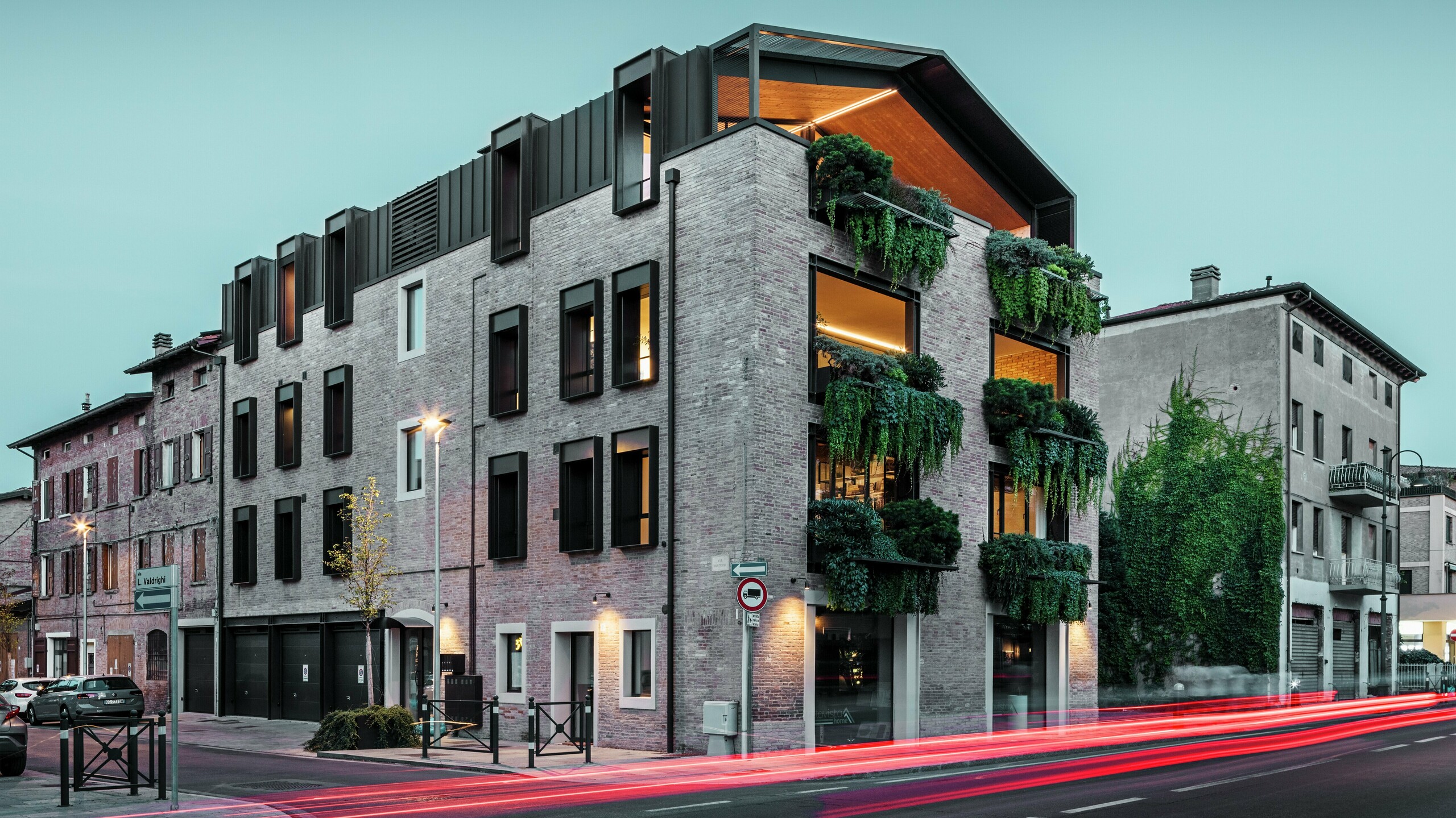 Zrównoważony budynek mieszkalno-usługowy we Włoszech z dachem aluminiowym PREFALZ w kolorze P.10 orzechowy brąz, zieloną fasadą z cegły i nowoczesną architekturą w wieczornej atmosferze przy świecących latarniach ulicznych.