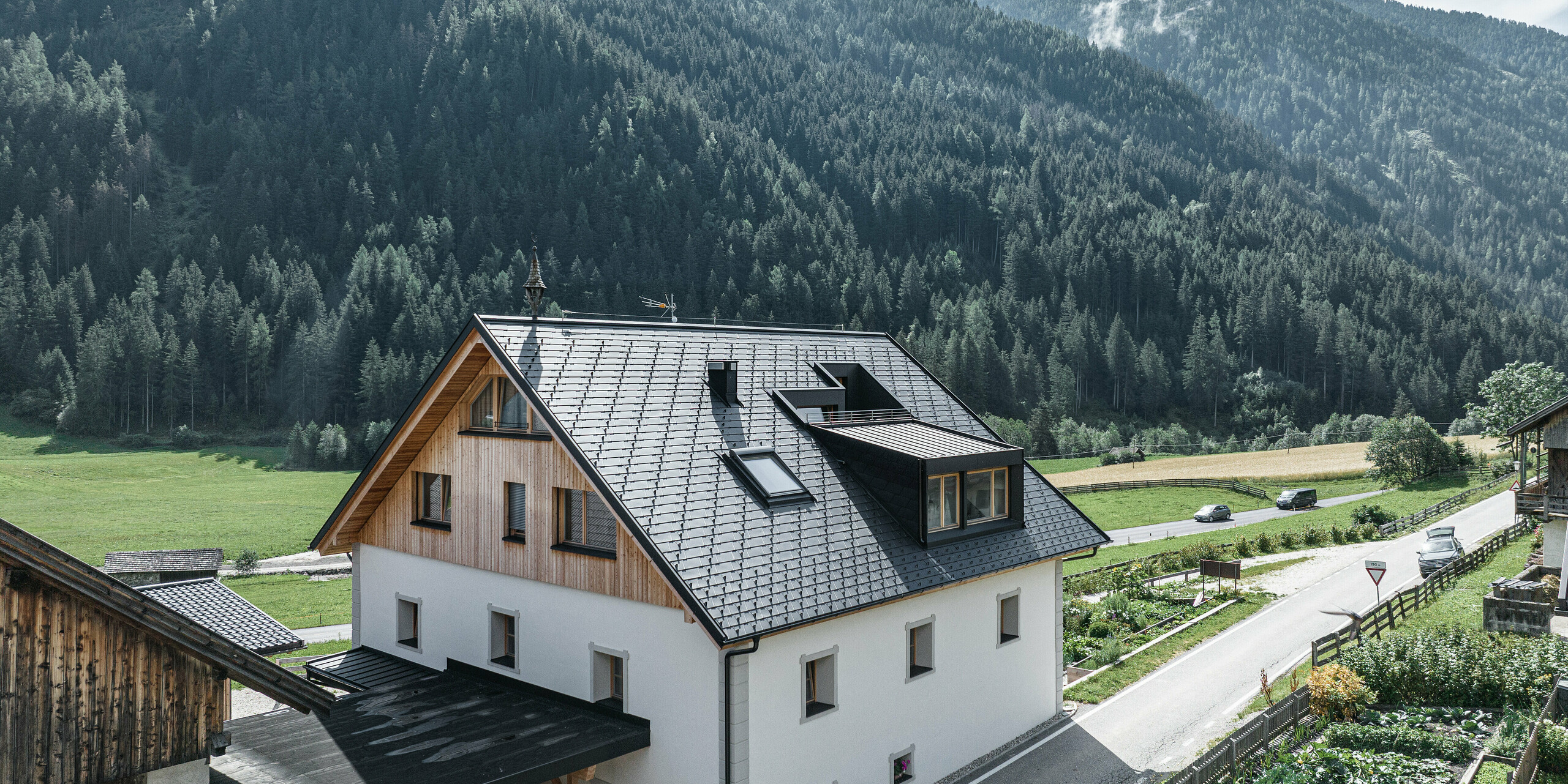 Perspektivische Ansicht des Ausserwegerhofs in Antholz, Südtirol, mit seinem modernen PREFA-Dach aus anthrazitfarbenen DS.19-Schindeln. Die weiße Fassade des Gebäudes hebt sich vom dichten Nadelwald und dem ruhigen Straßenbild ab. Die Giebel des Gebäudes sind mit Holz verkleidet und bilden einen harmonischen Übergang zum Aluminiumdach. Aus dem Giebeldach ragt auf beiden Seiten eine Gaube heraus. Unterhalb der Gauben befinden sich großzügige Fenster, die eine gute Aussicht ermöglichen.  Übersetzt mit DeepL.com (kostenlose Version)