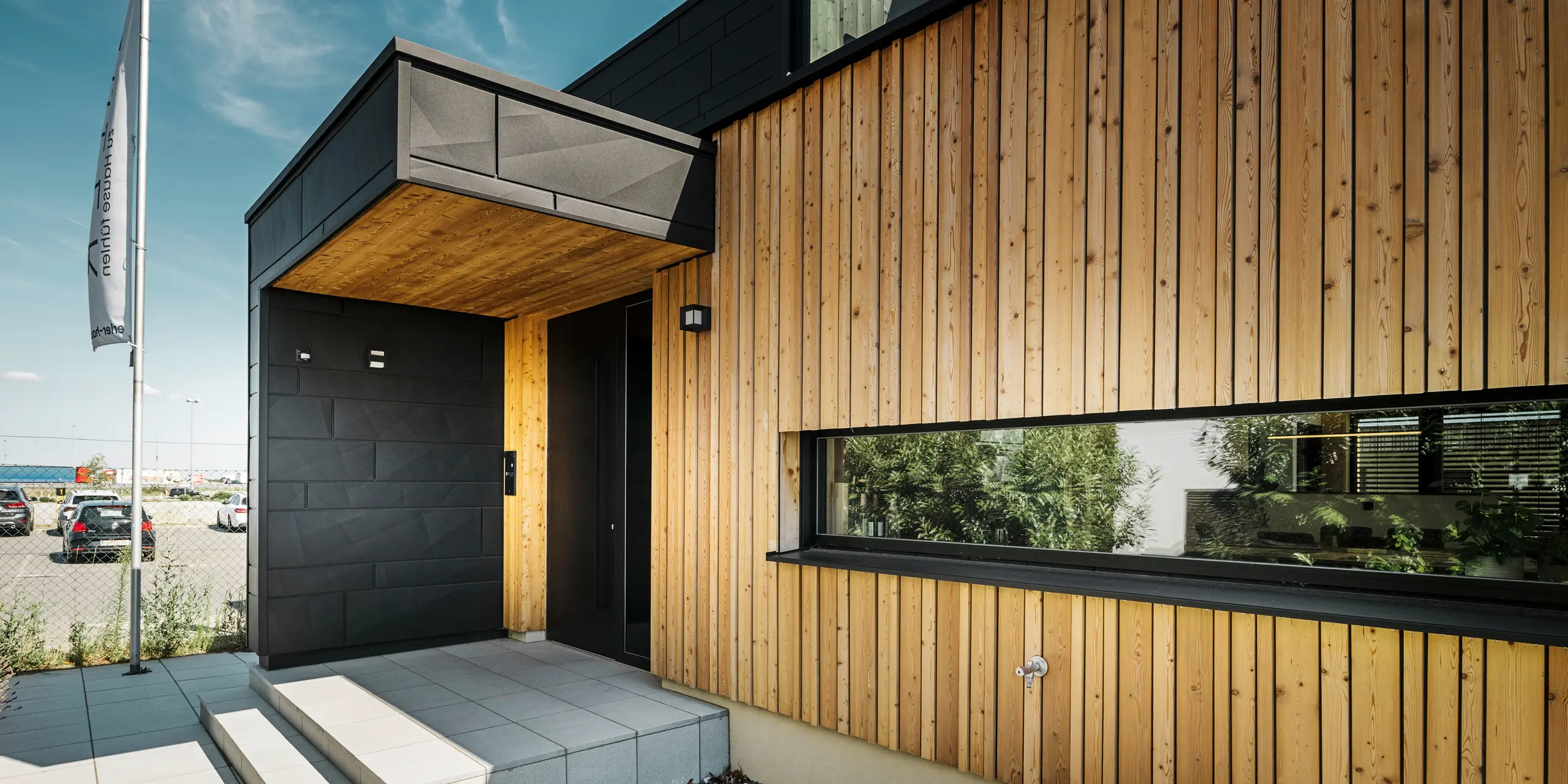 Boczne wejście do domu pokazowego Autark w Centrum domów prefabrykowanych Blaue Lagune w Vösendorf, z nowoczesną fasadą wykonaną z naturalnego drewna i czarnych paneli Siding.X PREFA. Zdjęcie przedstawia eleganckie połączenie okładziny drewnianej i aluminium w wysokiej jakości powłoce P.10 czarny.