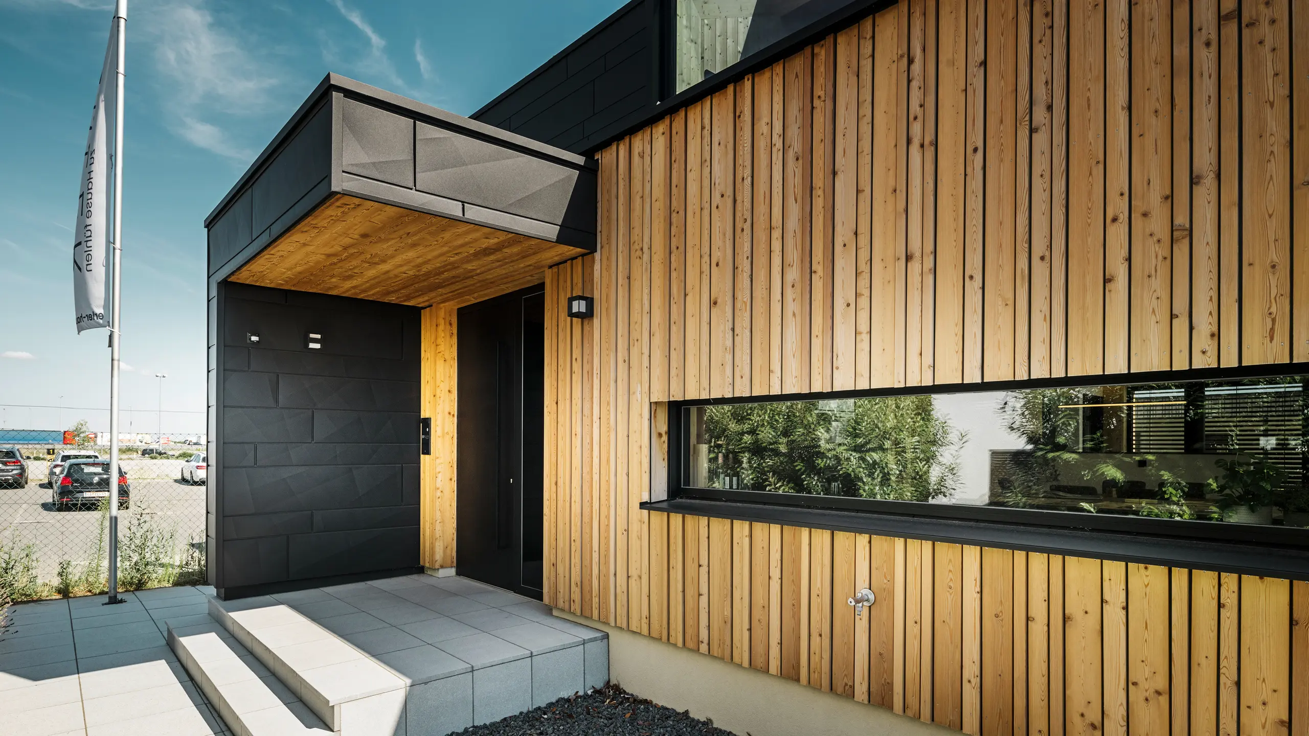 Boczne wejście do domu pokazowego Autark w Centrum domów prefabrykowanych Blaue Lagune w Vösendorf, z nowoczesną fasadą wykonaną z naturalnego drewna i czarnych paneli Siding.X PREFA. Zdjęcie przedstawia eleganckie połączenie okładziny drewnianej i aluminium w wysokiej jakości powłoce P.10 czarny.