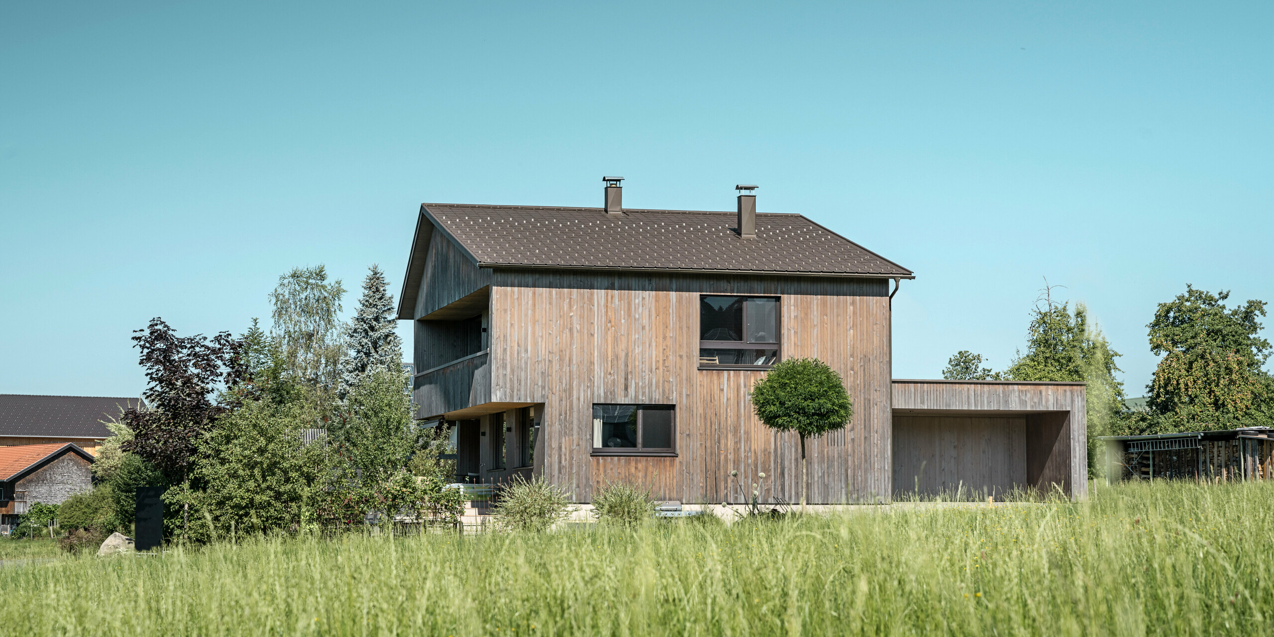 Modernes Einfamilienhaus in Krumbach mit einer nachhaltigen Holzfassade und einem robusten Aluminiumdach aus PREFA Dachplatten in P.10 Braun. Die natürlichen Farben und Materialien des Hauses fügen sich nahtlos in die ländliche Umgebung ein, während das PREFA Dachsystem robusten Schutz und eine ästhetische Komponente bietet. Rund um das Gebäude ist eine natürliche Landschaft mit etlichen Bäumen und einer grünen Wiese im Vordergrund zu erkennen. Das Dach des Gebäudes hebt sich mit seinem kräftigen Braunton von der verblasten Holzfassade ab und betont zusätzlich die Beständigkeit von der Farbqualität P.10.