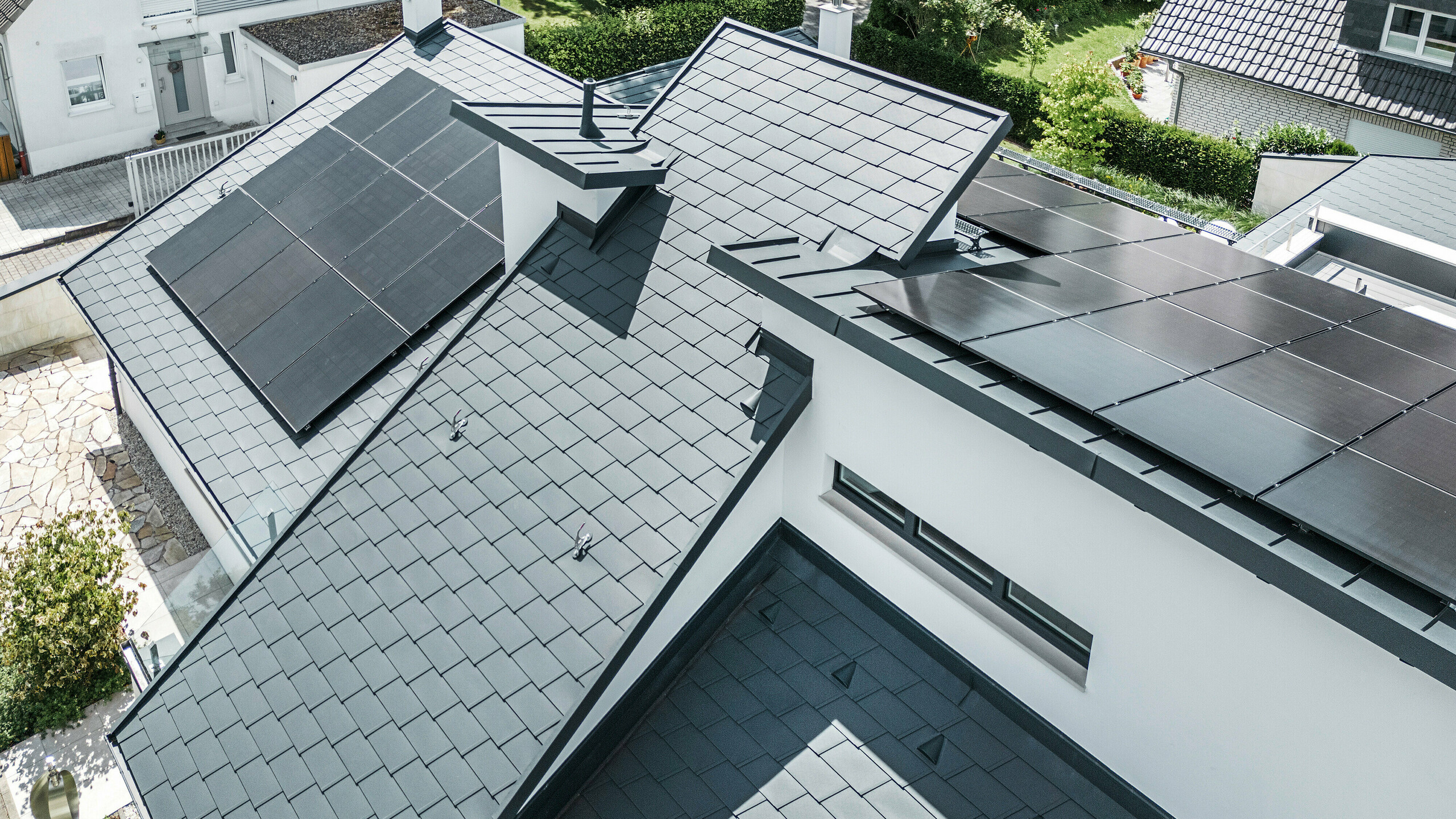 Widok z lotu ptaka na różne poziomy na dachu nowoczesnego domu jednorodzinnego w Dortmundzie, który został wyposażony w gonty dachowe PREFA DS.19 i PREFALZ w kolorze P.10 antracyt oraz system fotowoltaiczny na dużą skalę.
