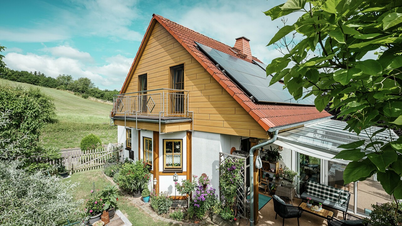 To zdjęcie przedstawia urokliwy dom jednorodzinny w Blieskastel, którego świeżo odnowiony szczyt wykończony sidingiem PREFA w kolorze dąb naturalny od razu przyciąga wzrok. Ciepła estetyka drewna uzyskana dzięki trwałemu aluminium harmonijnie komponuje się z czerwonym dachem z dachówki oraz przestronnym ogrodem zimowym. Otoczony bujną zielenią i starannie zaprojektowanymi rabatami kwiatowymi tworzy idylliczną, pełną życia scenę. Solidny remont elewacji z PREFA nadaje domowi naturalny wygląd, ale też zachęca odpornością na warunki atmosferyczne, trwałością koloru i długim, bezobsługowym użytkowaniem.