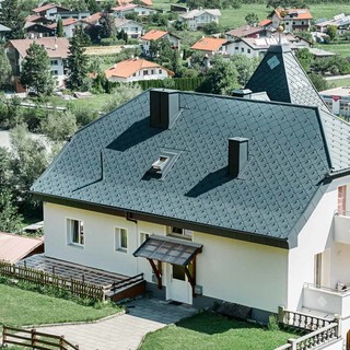 Klasyczny dach szczytowy z dwoma oknami dachowymi i wieloma dodatkowymi oknami, kryty dachówką rombową PREFA 44 x 44 w kolorze jasnoszarym.