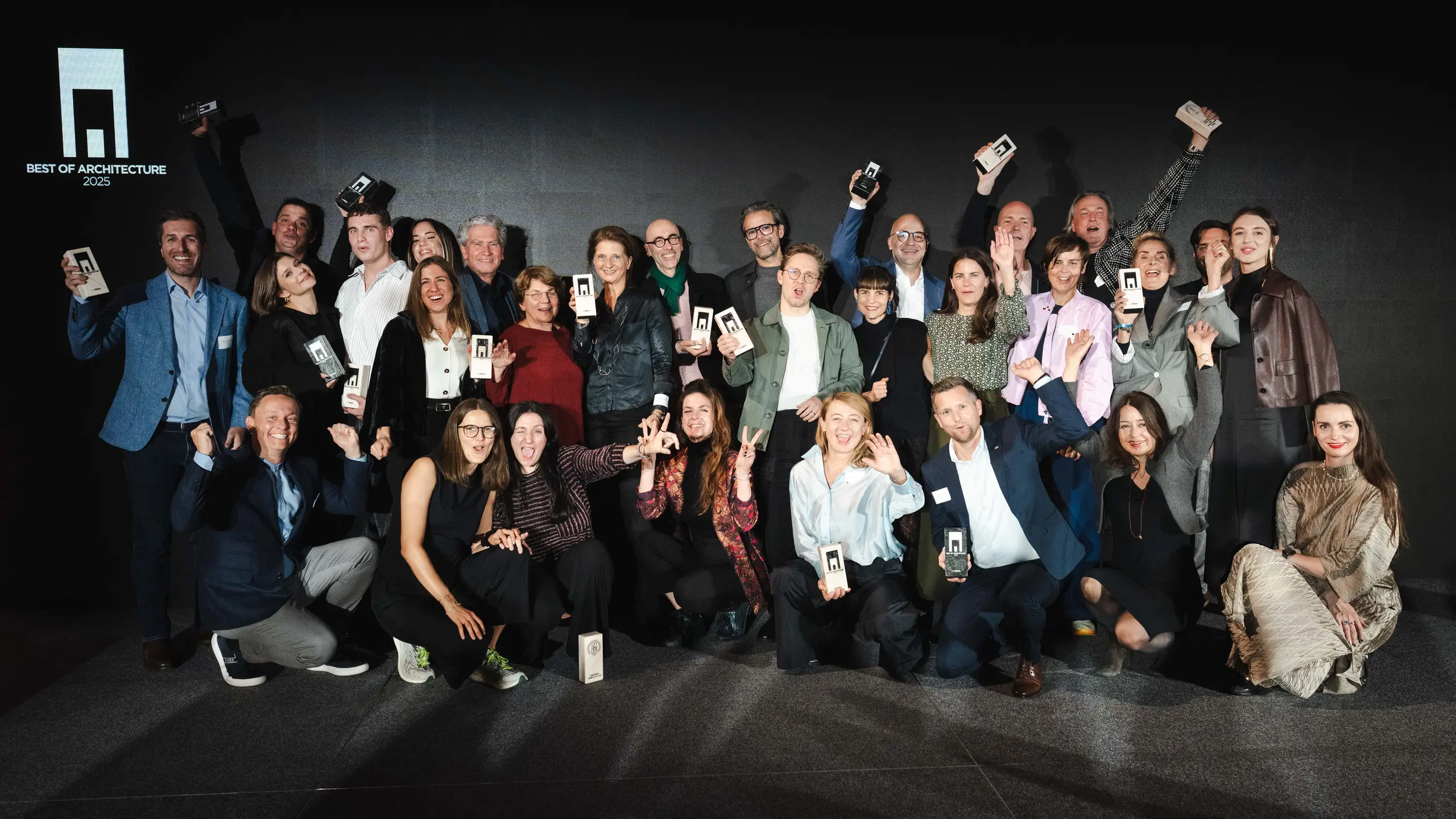 Zdjęcie grupowe zwycięzców podczas ceremonii wręczenia nagród „Budynek mieszkalny roku 2025”: zwycięzcy stoją przed elegancką czarną sceną z logo „Best of Architecture” i dumnie trzymają w rękach swoje nagrody. Dynamiczne pozy, nowoczesne ubrania i efektowne oświetlenie podkreślają znaczenie wydarzenia, a scena symbolizuje innowacyjność i doskonałość architektoniczną.