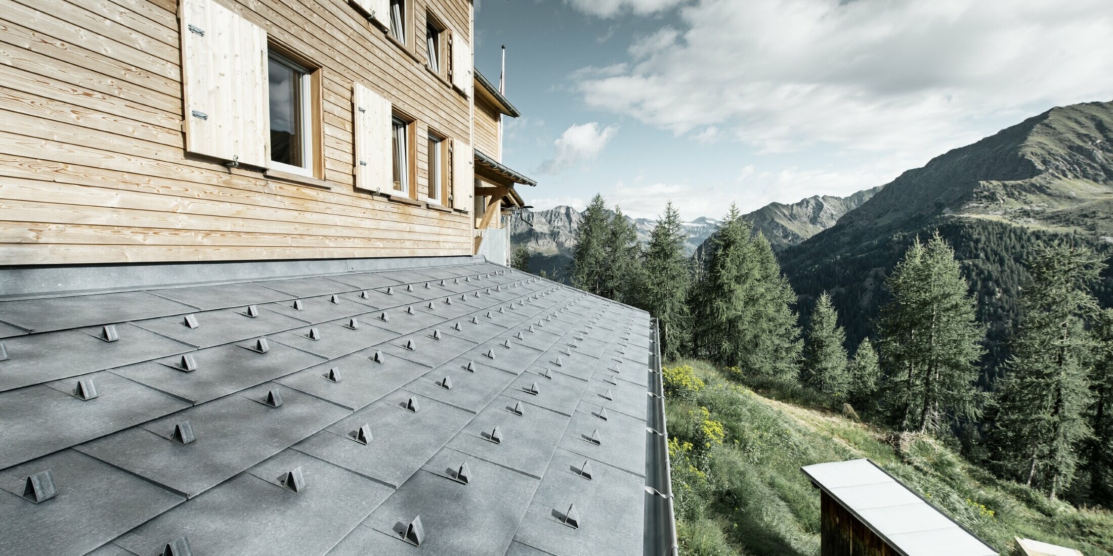 Zbliżenie zadaszenia nad wejściem do drewnianej chaty. Zadaszenie pokryte jest panelami aluminiowymi PREFA FX.12 w kolorze szary kamienny ze stoperami śniegu. Rynna dachowa podtrzymywana jest przez wsporniki.