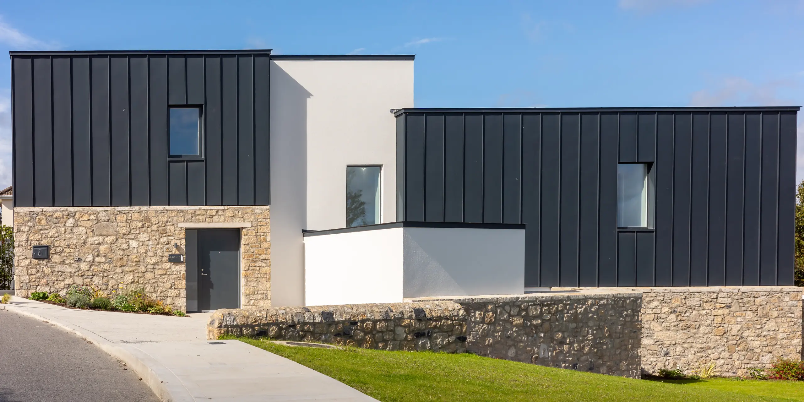 Modernes Wohnhaus in Watson Place mit klaren Linien, Natursteinmauer und markanter PREFALZ Dach‑ und Fassadenverkleidung in P.10 Anthrazit. Die glatten Aluminiumflächen setzen elegante Kontraste zu Stein und Putz und bieten langlebigen Schutz vor Witterung. Das Zusammenspiel der Materialien verleiht dem Gebäude eine hochwertige, zeitlose Erscheinung.