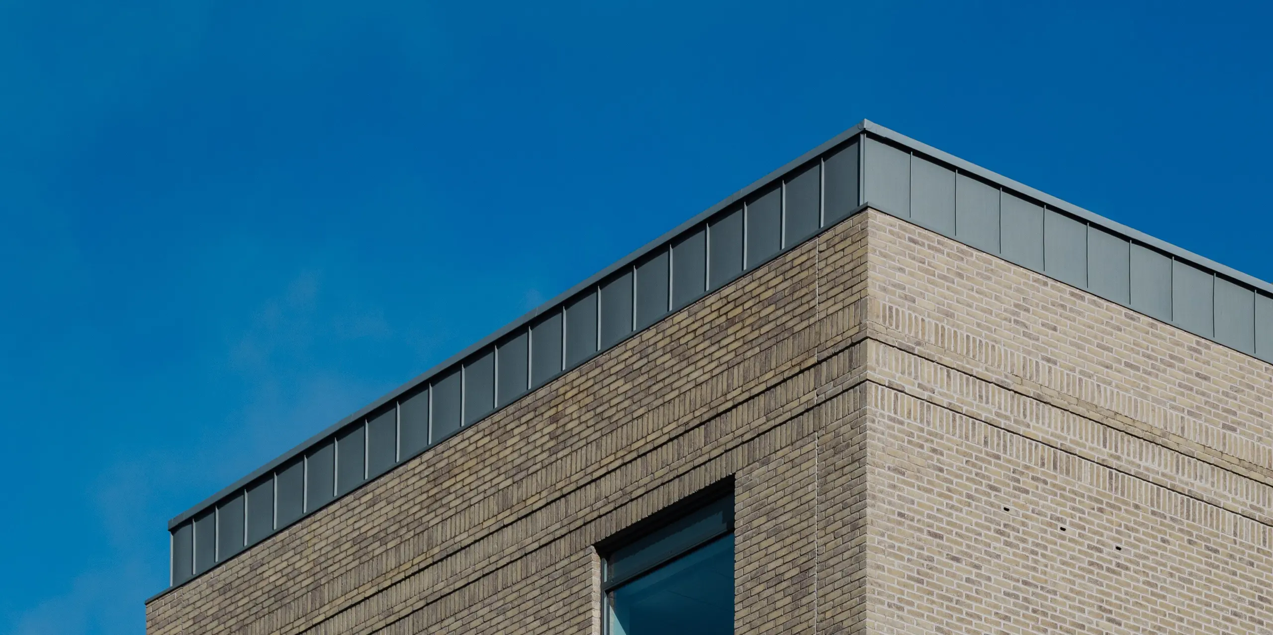 Das Northern Regional College in Ballymena zeigt sich als markanter Bildungsbau mit klaren Linien und hochwertiger Materialität. Die Fassade aus hellem Klinker wird von vertikal verlegten PREFALZ Aluminium-Stehfalzbahnen in Patinagrau gekrönt, die als visueller Eyecatcher dienen. Das langlebige PREFA Aluminium überzeugt durch Witterungsbeständigkeit, geringes Gewicht und elegante Optik. Der ruhige Grauton setzt einen spannenden Kontrast zu Glasflächen und Himmel und verleiht dem Gebäude zeitlose architektonische Präsenz.