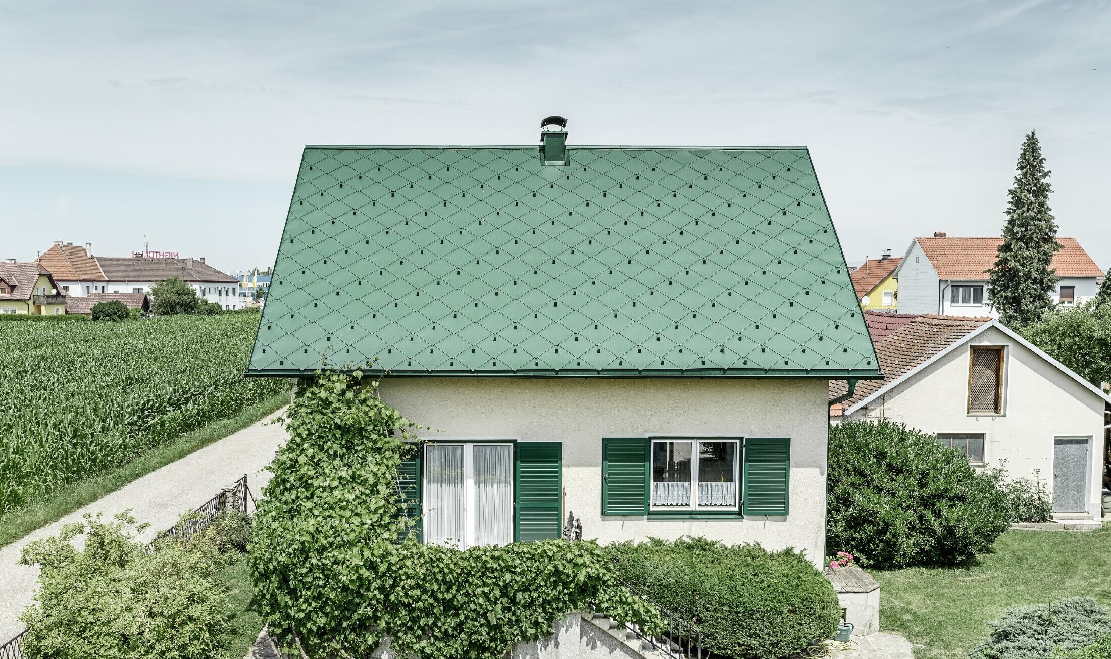 Klasyczny dom jednorodzinny z dachem dwuspadowym, pokryty aluminium w kolorze zieleni mchu z zielonymi okiennicami. Dach pokryto dachówką romb PREFA 44 × 44 w kolorze P.10 zieleń mchu.
