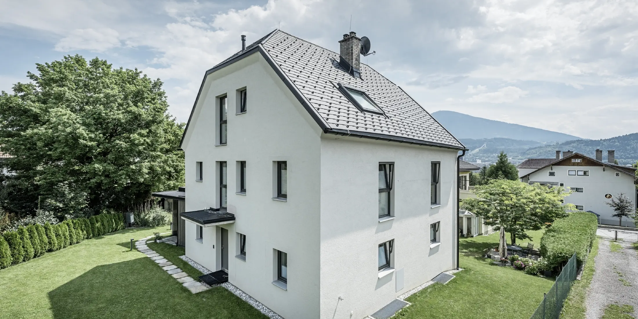 Nowoczesny trzypiętrowy dom jednorodzinny z jasną elewacją tynkową i grafitowym dachem dwuspadowym z aluminium firmy PREFA. Budynek posiada kilka wąskich okien i system odprowadzania wody deszczowej w kolorze antracyt. Stoi on w zadbanym ogrodzie z trawnikiem, drzewami i żywopłotami, a w tle widać kolejne domy mieszkalne i górzysty krajobraz.