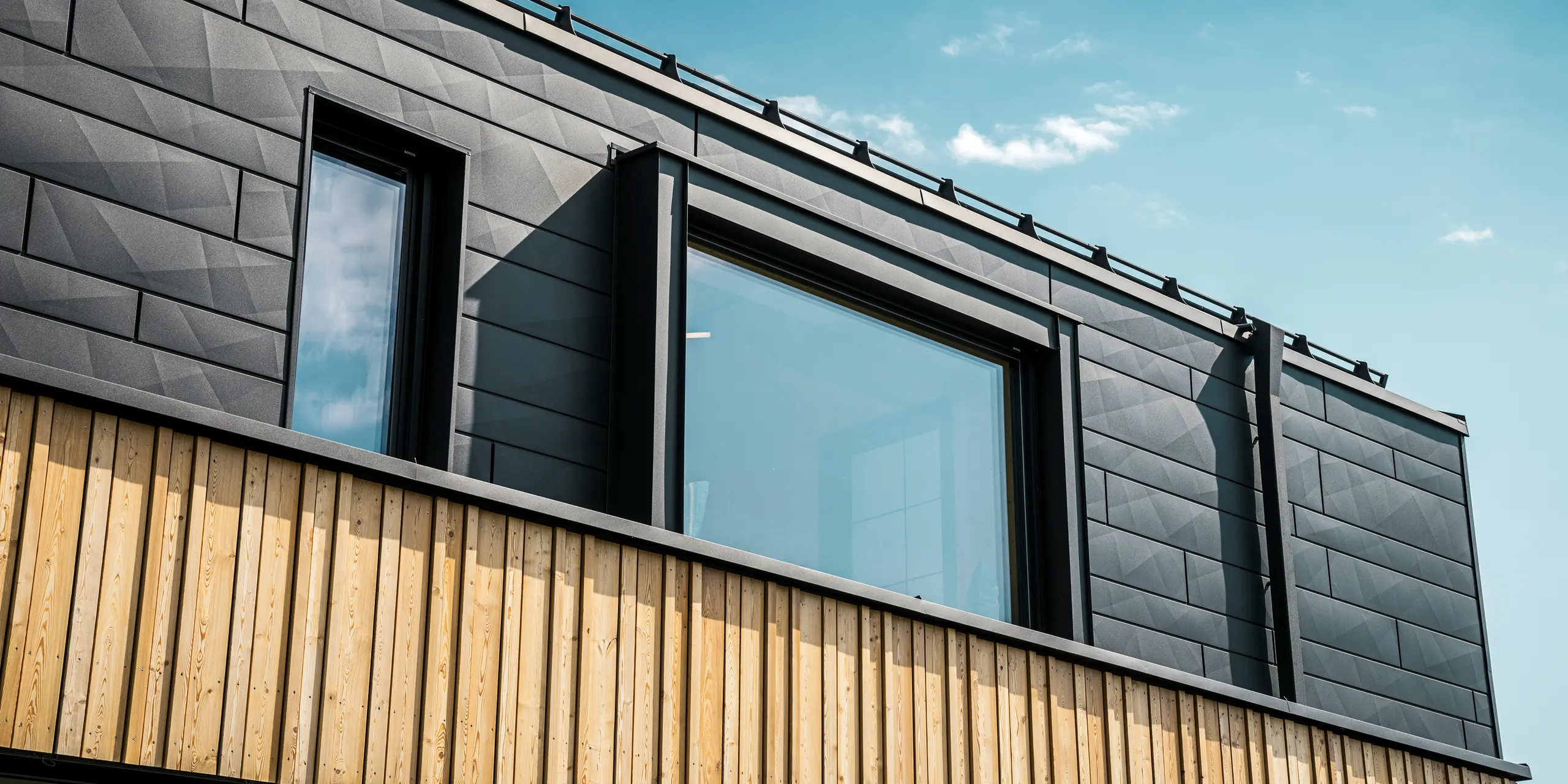Zbliżenie na górną kondygnację domu modelowego Autark w centrum domów prefabrykowanych Blaue Lagune w Vösendorfie (AT). Elewacja wykonana jest z paneli aluminiowych PREFA Siding.X w kolorze P.10 czarny, które wyróżniają się specjalnym wyglądem z różnymi kątami. Poniżej znajduje się stylowa okładzina drewniana, która stanowi harmonijny kontrast z ciemnym aluminium. Duże okna wpuszczają do wnętrza budynku dużo światła i podkreślają nowoczesny design.