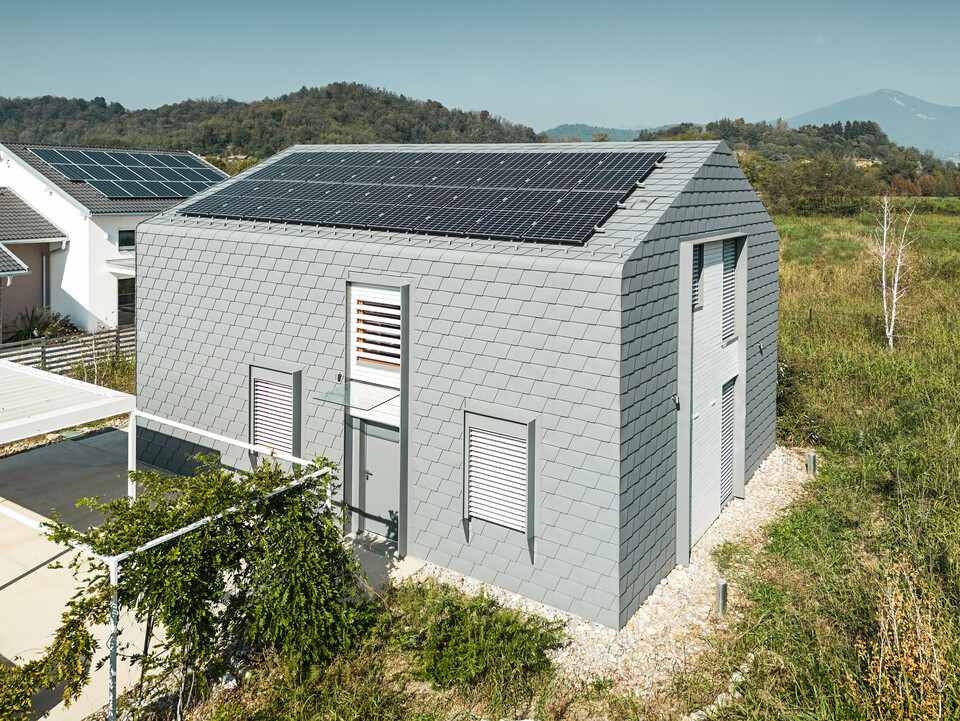Drohnenaufnahme eines modernen Gebäudes mit einer Hülle aus PREFA Dachschindeln und Wandschindeln in der Farbe P.10 Hellgrau. Die Gebäudehülle zeichnet sich durch ein gleichmäßiges, schuppenartiges Muster aus, das eine zeitgemäße Optik bietet. Am Dach des Einfamilienhauses ist eine Photovoltaikanlage zu sehen. Im Hintergrund ist ein Haus ebenfalls mit einer Photovoltaikanlage ausgestattet wurden, was den Fokus auf Nachhaltigkeit und moderne Architektur legt.