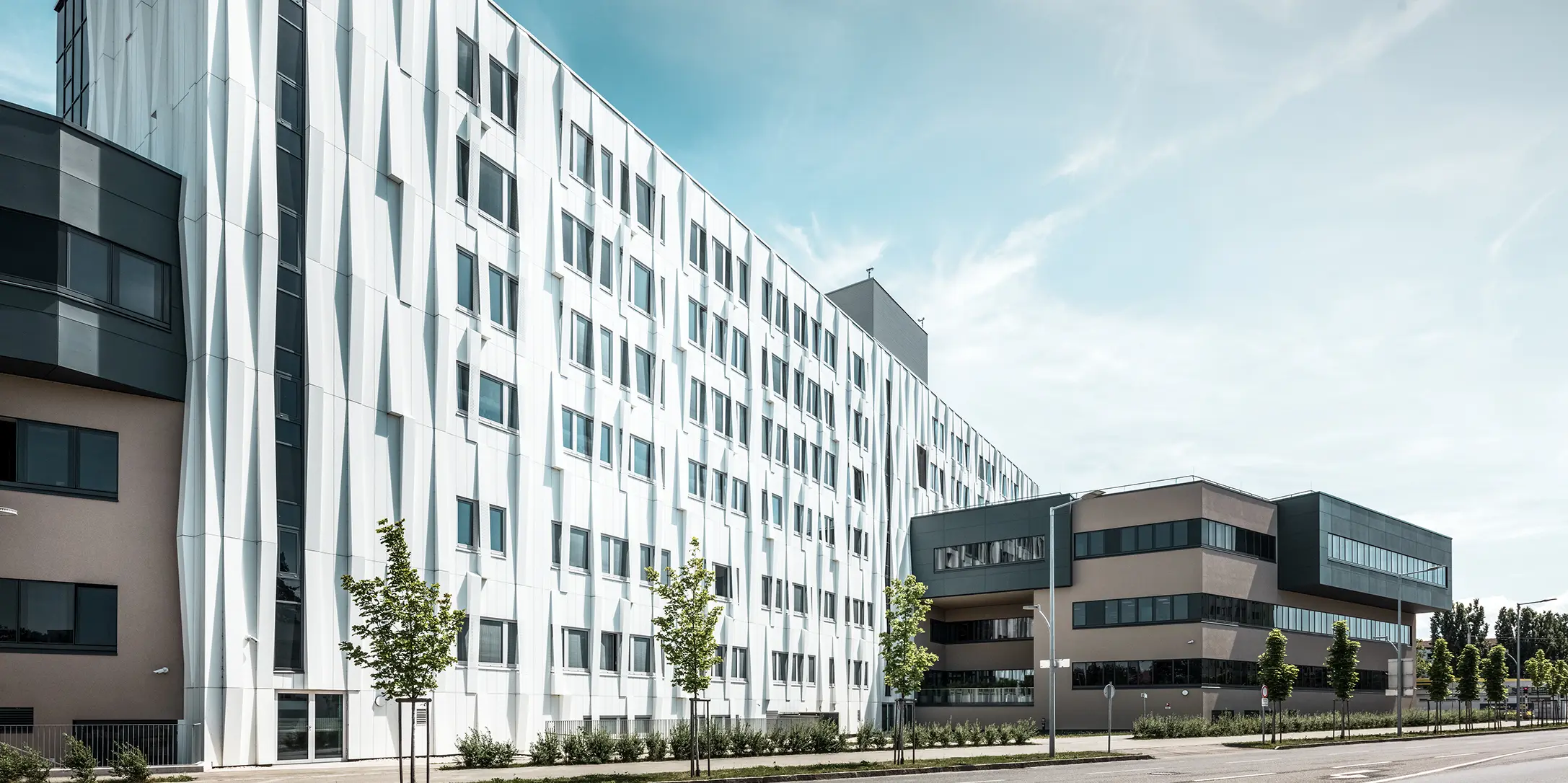 Widok z boku kliniki uniwersyteckiej w St. Pölten w Dolnej Austrii. Solidna aluminiowa fasada sprawia, że budynek przyciąga wzrok w mieście. Na pierwszym planie widać ulicę z kilkoma drzewami.