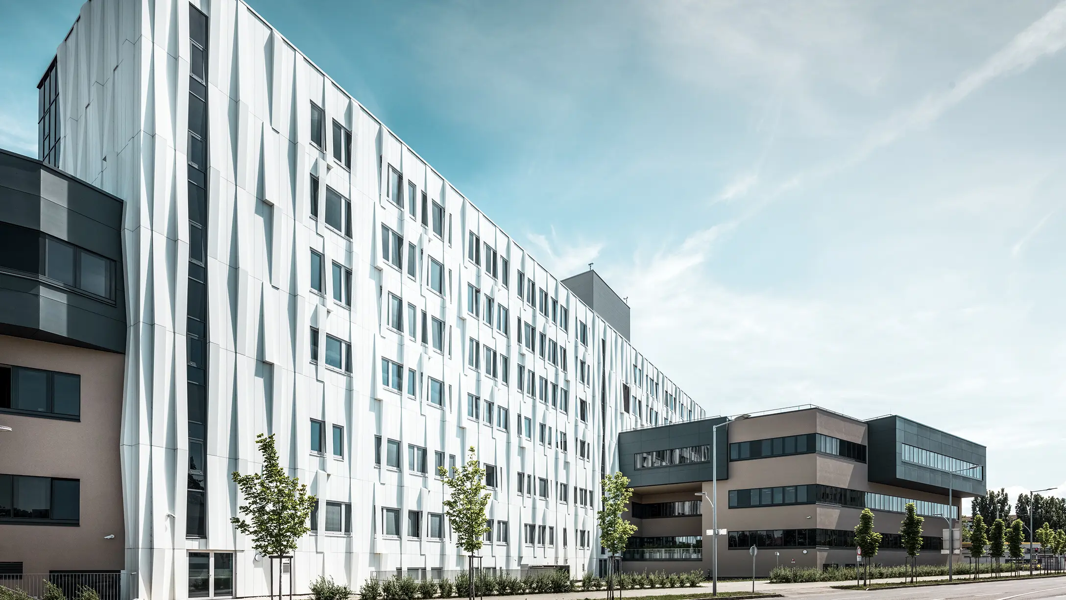 Widok z boku kliniki uniwersyteckiej w St. Pölten w Dolnej Austrii. Solidna aluminiowa fasada sprawia, że budynek przyciąga wzrok w mieście. Na pierwszym planie widać ulicę z kilkoma drzewami.