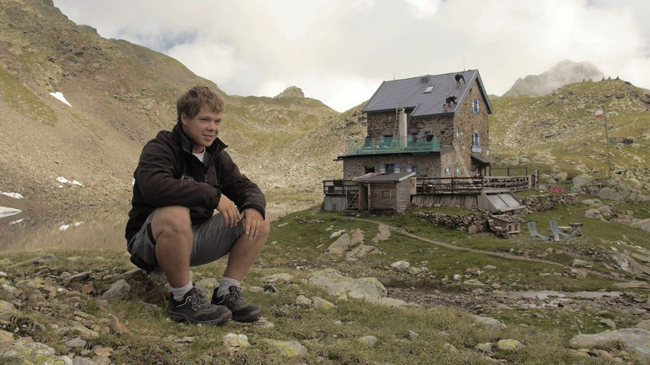 Praktykant Jakob Schöttl siedzi na kamieniu. W tle widoczne jest schronisko „Flagger Schartenhütte”, pokryte dachówką klasyczną PREFA.